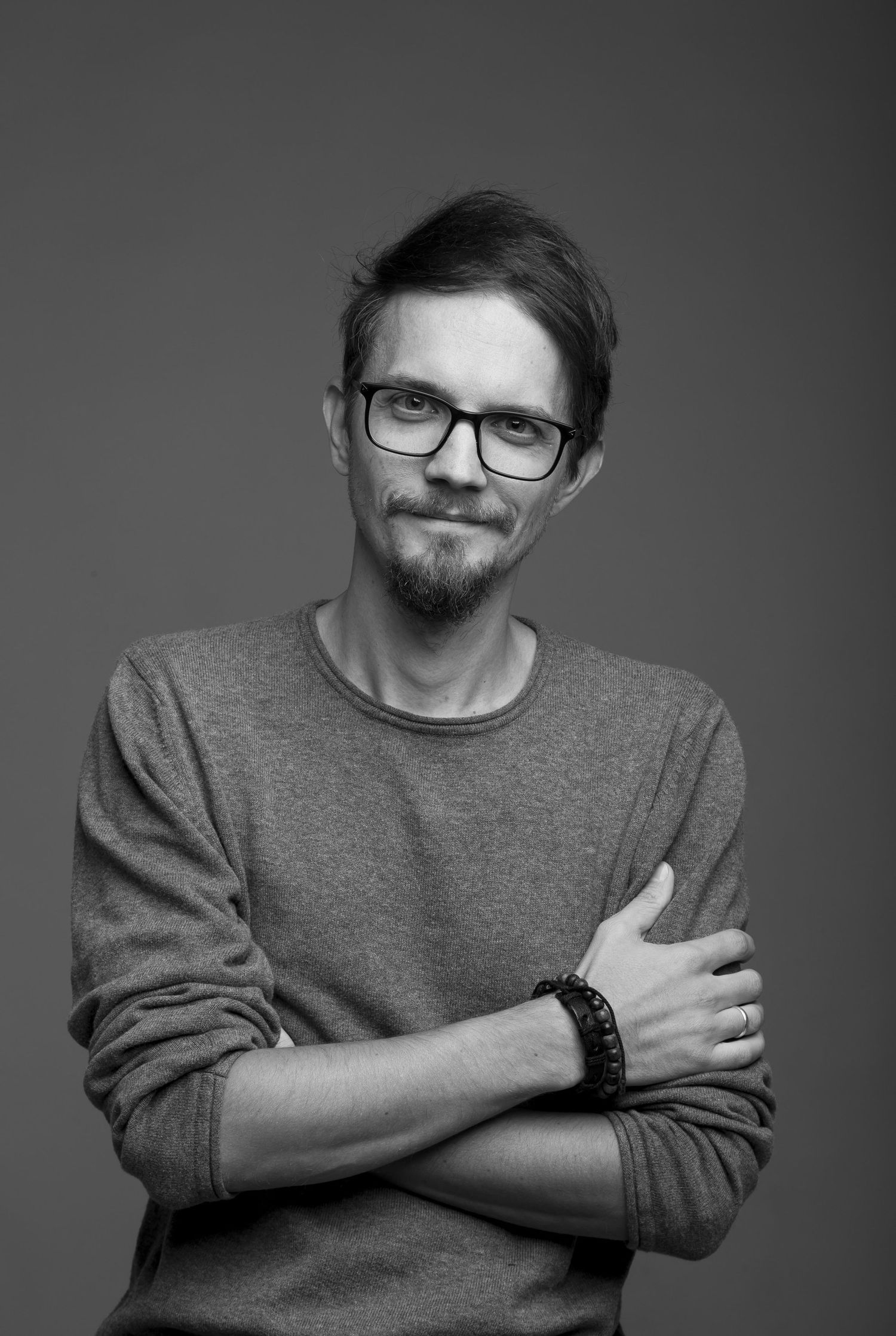 Black and white portrait of man in gray sweater and glasses with arms crossed, smiling slightly.