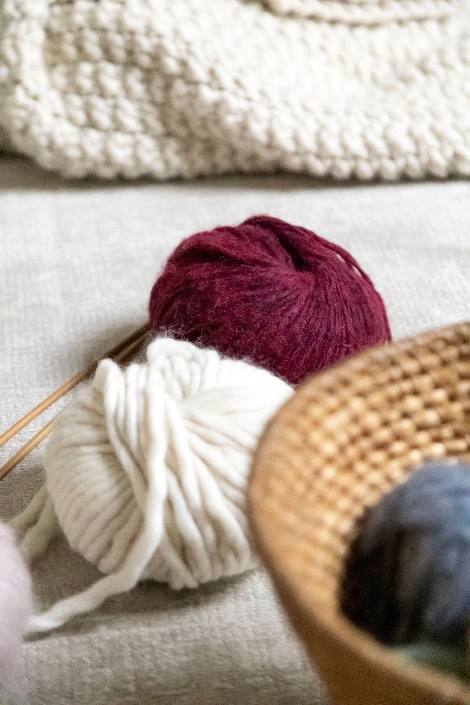 Burgundy and white yarn balls with knitting needles beside a wicker basket on linen.