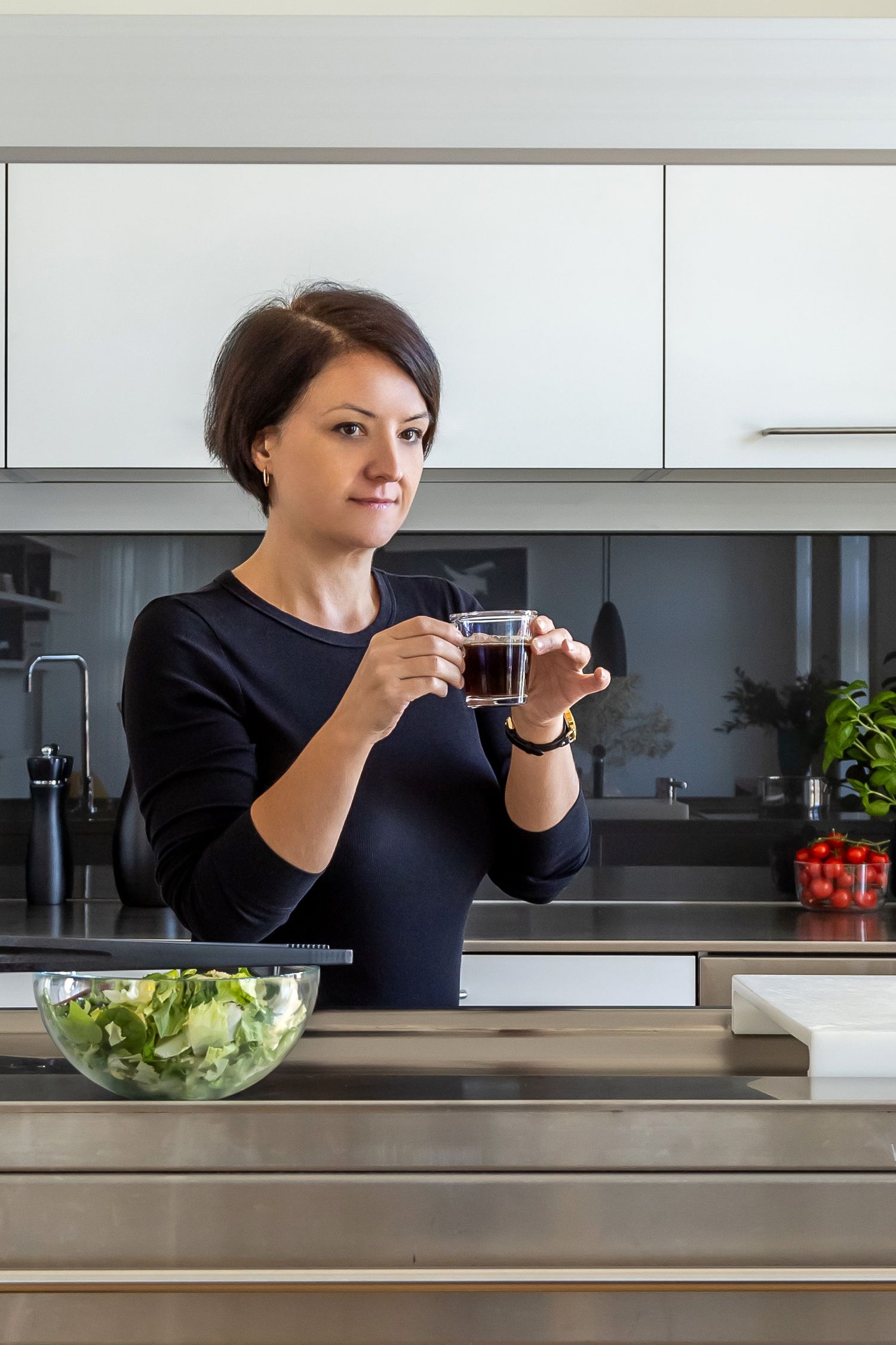 Woman in black top holding espresso glass at modern kitchen counter with fresh salad bowl.