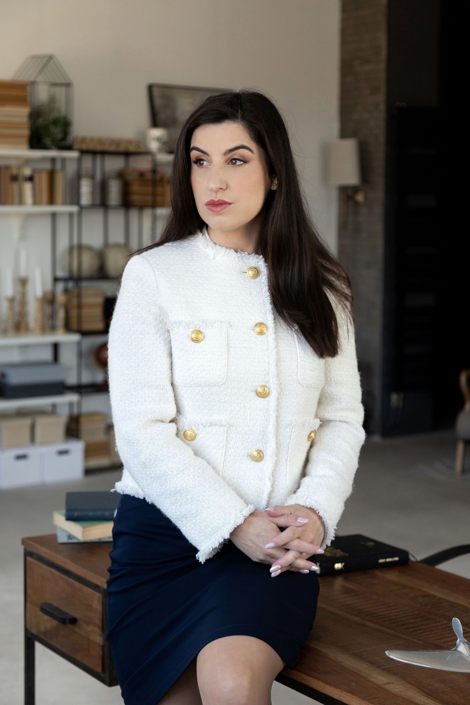 Professional woman in white tweed jacket and navy skirt sits on desk in modern office setting.