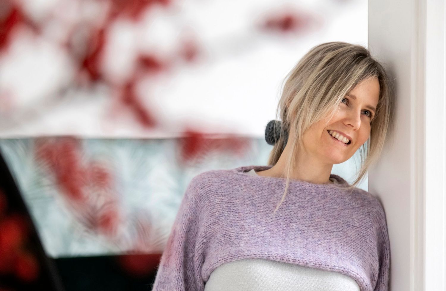 Smiling blonde woman in a lavender knit sweater leaning against a white wall relaxing.
