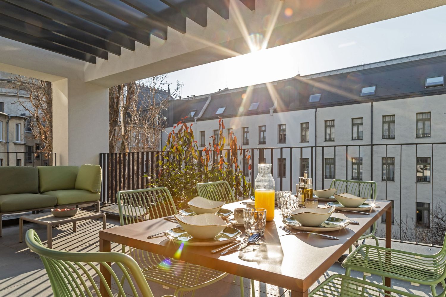 Sunny rooftop terrace with wooden dining table, green chairs, and city building views in the background.