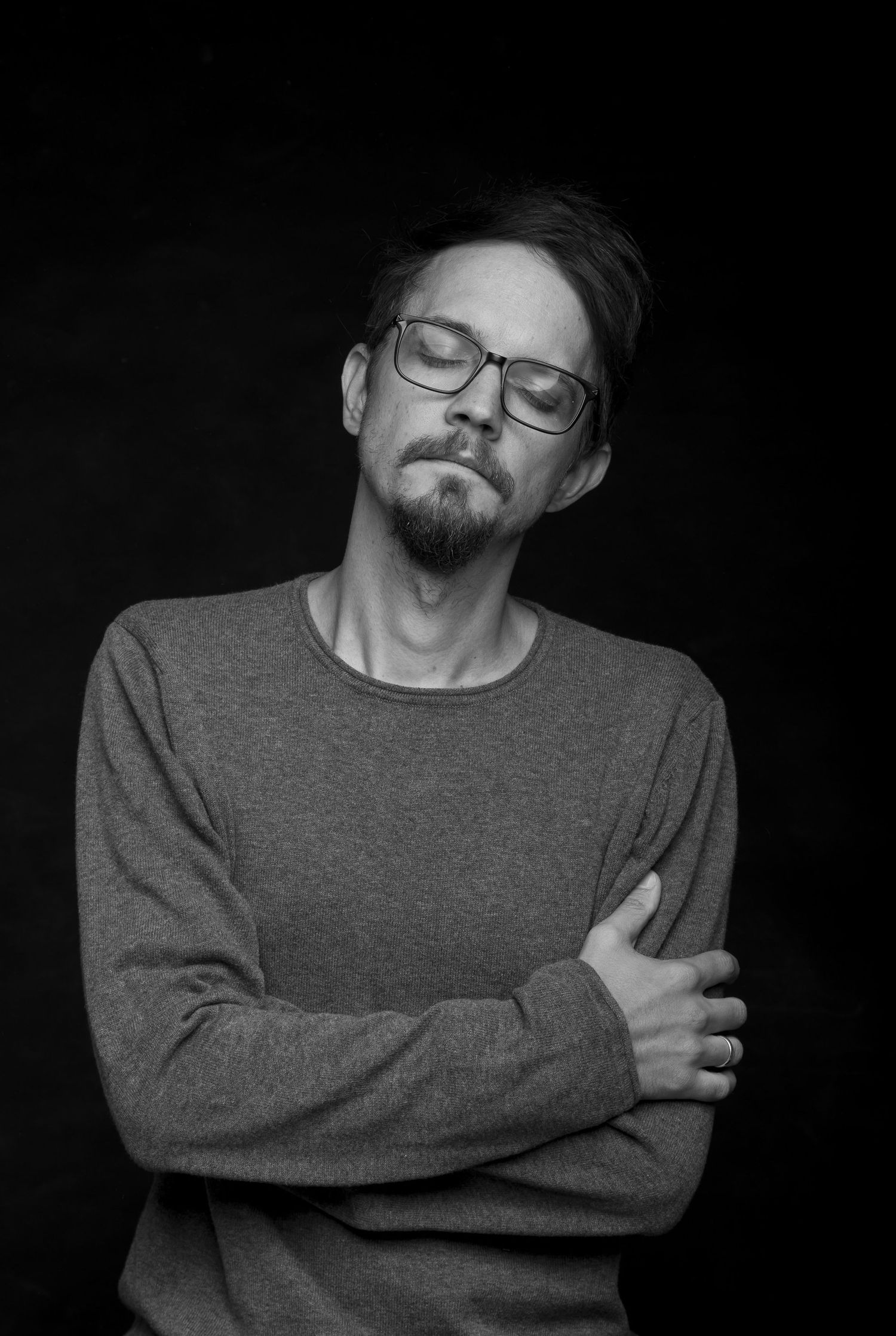 Black and white close-up portrait of bearded man in gray sweater, glasses, arms crossed, eyes closed.