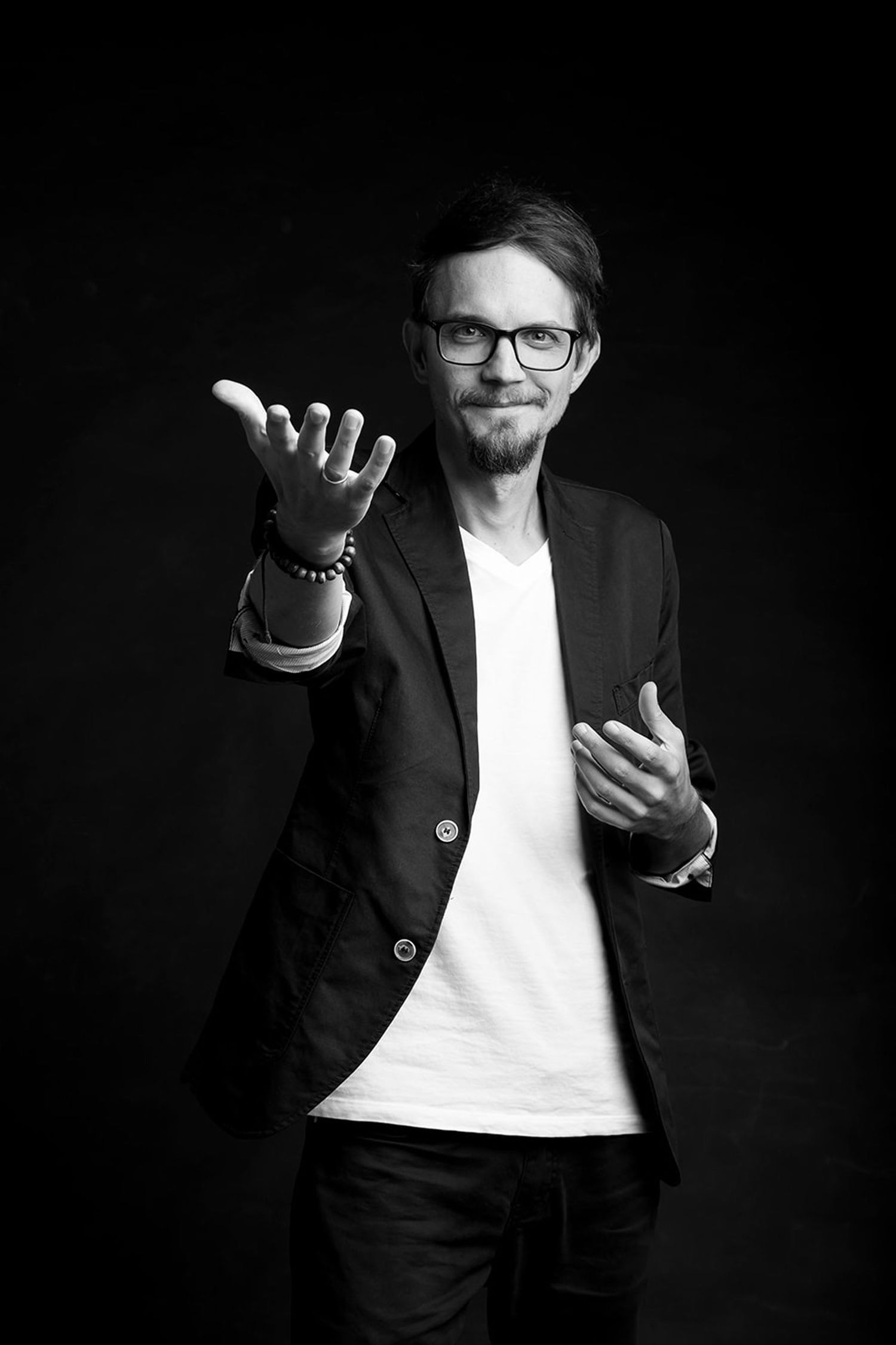 Black and white photo of man in blazer and glasses reaching toward camera with open hand gesture.