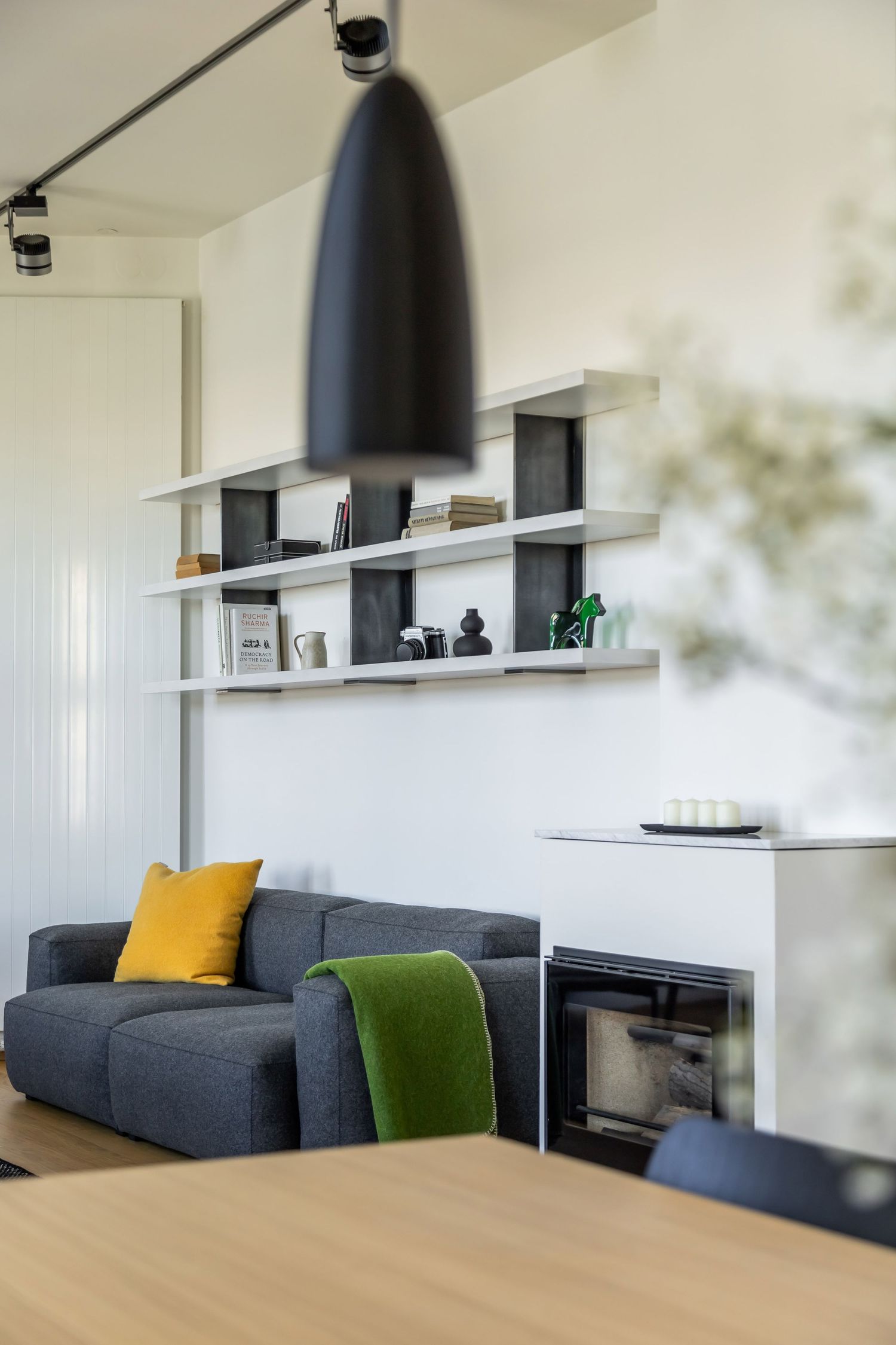 Modern living room with a dark gray sofa, yellow and green cushions, wall-mounted shelving unit, and black pendant light.