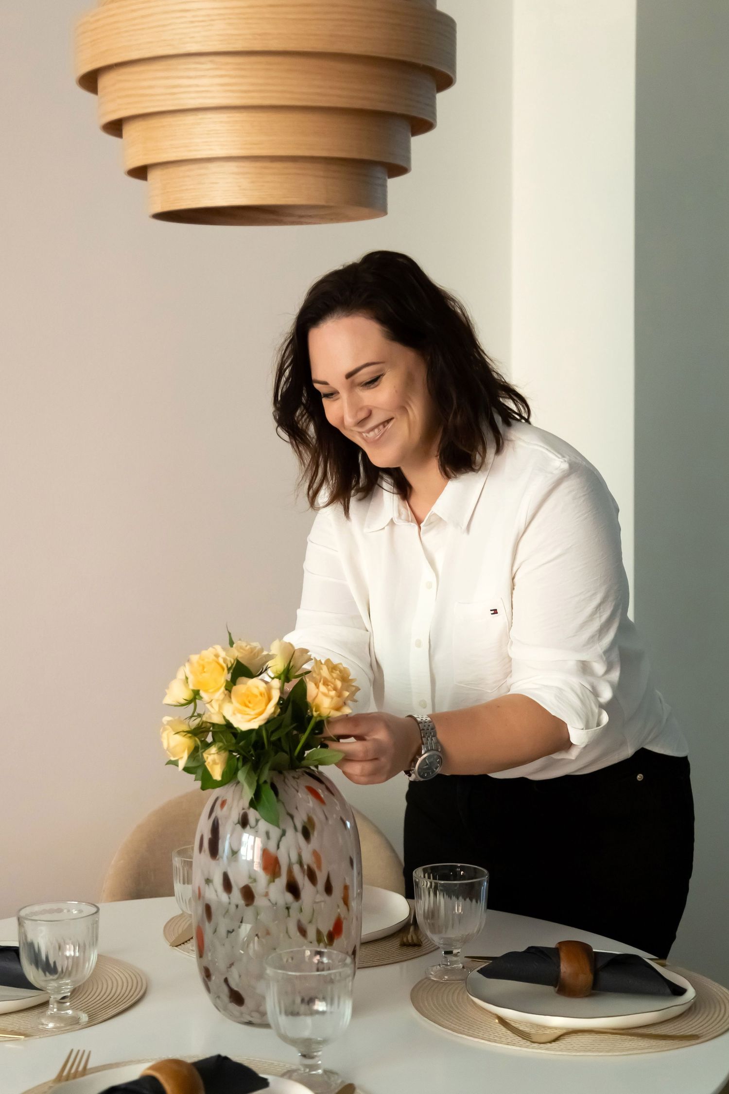 Cheerful woman in white blouse arranging fresh yellow roses in a decorative vase on a styled dining table.