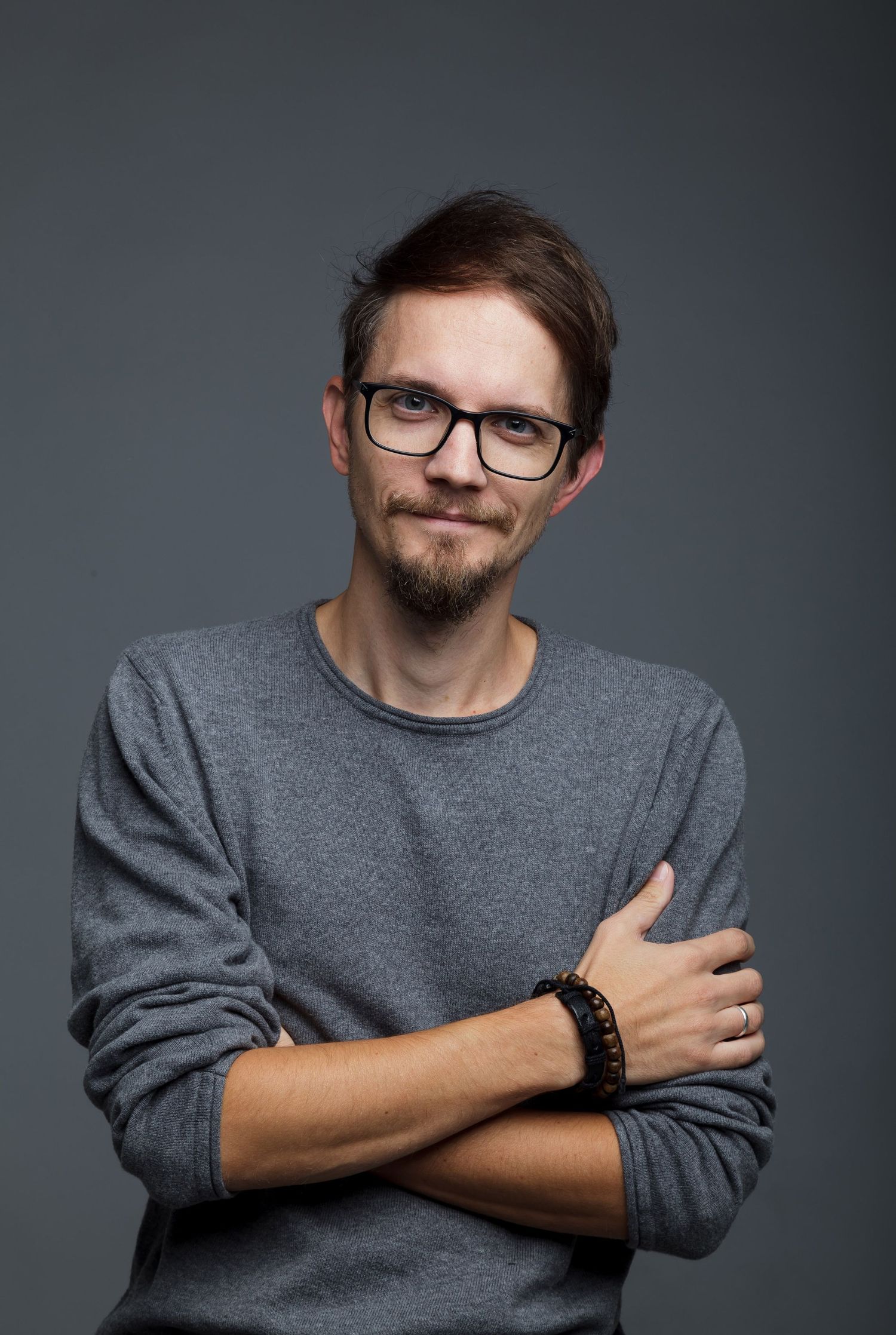 Young man with glasses and beard wearing gray sweater, arms crossed, posing against dark gray background.