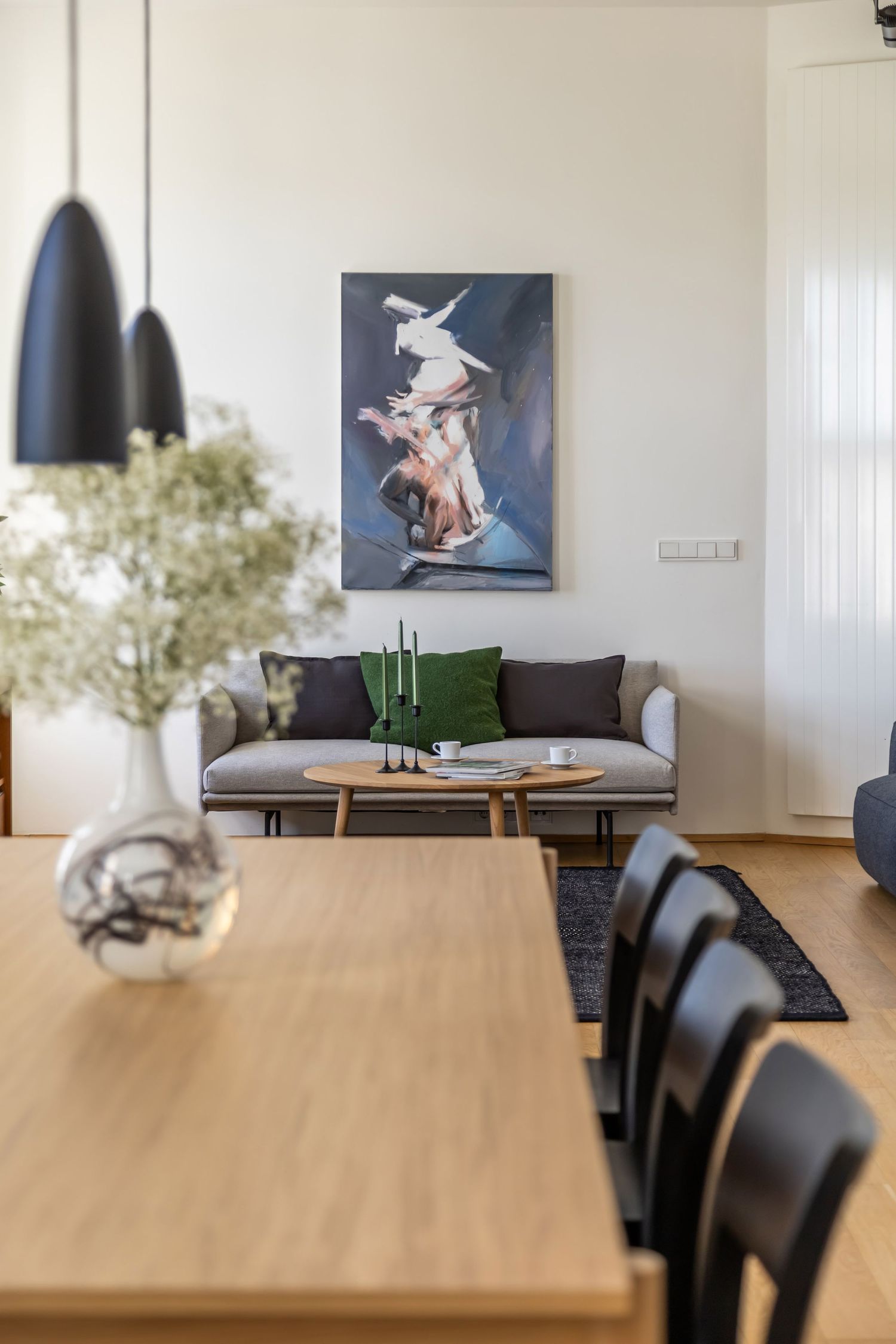Open-plan living and dining area with a gray sofa, green pillows, dramatic figurative painting, and black pendant lights.