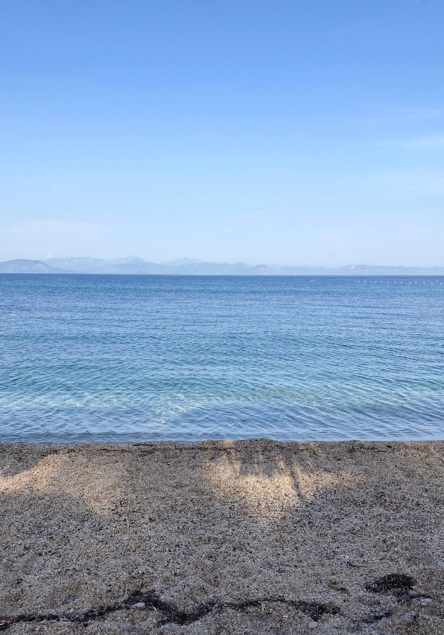 Serene beach scene with calm blue ocean waters, sandy shore, and distant mountains under a clear sky.