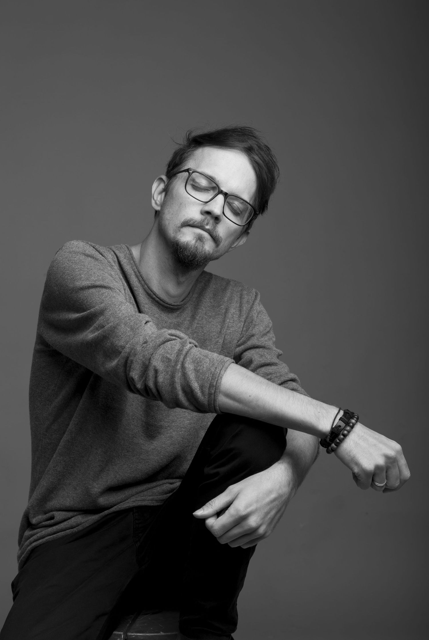 Black and white portrait of a young man with glasses and beard wearing a sweater and bracelet, posing against gray background.