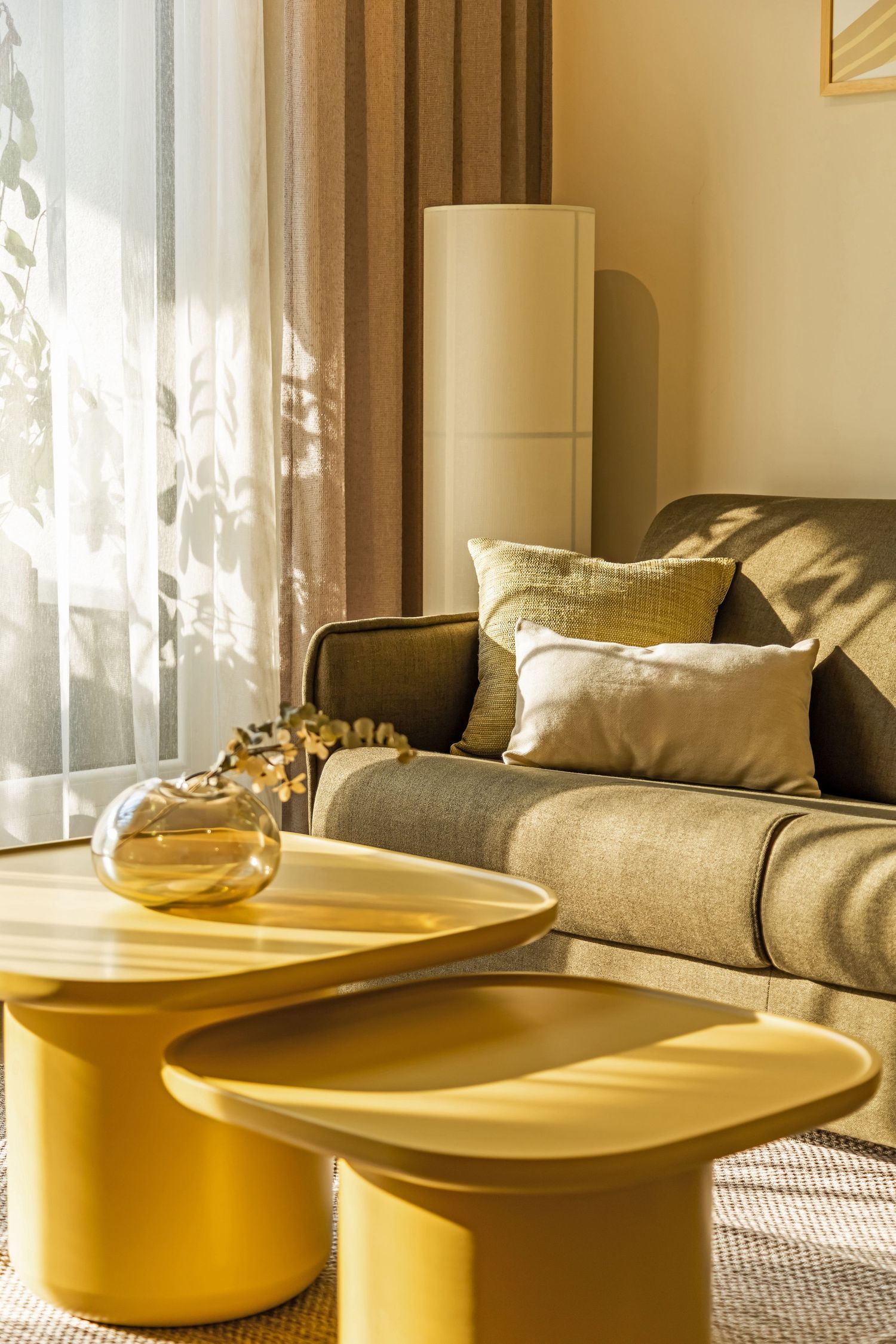Bright living room with yellow coffee tables, gray sofa, decorative pillows, and sheer curtains in warm sunlight.