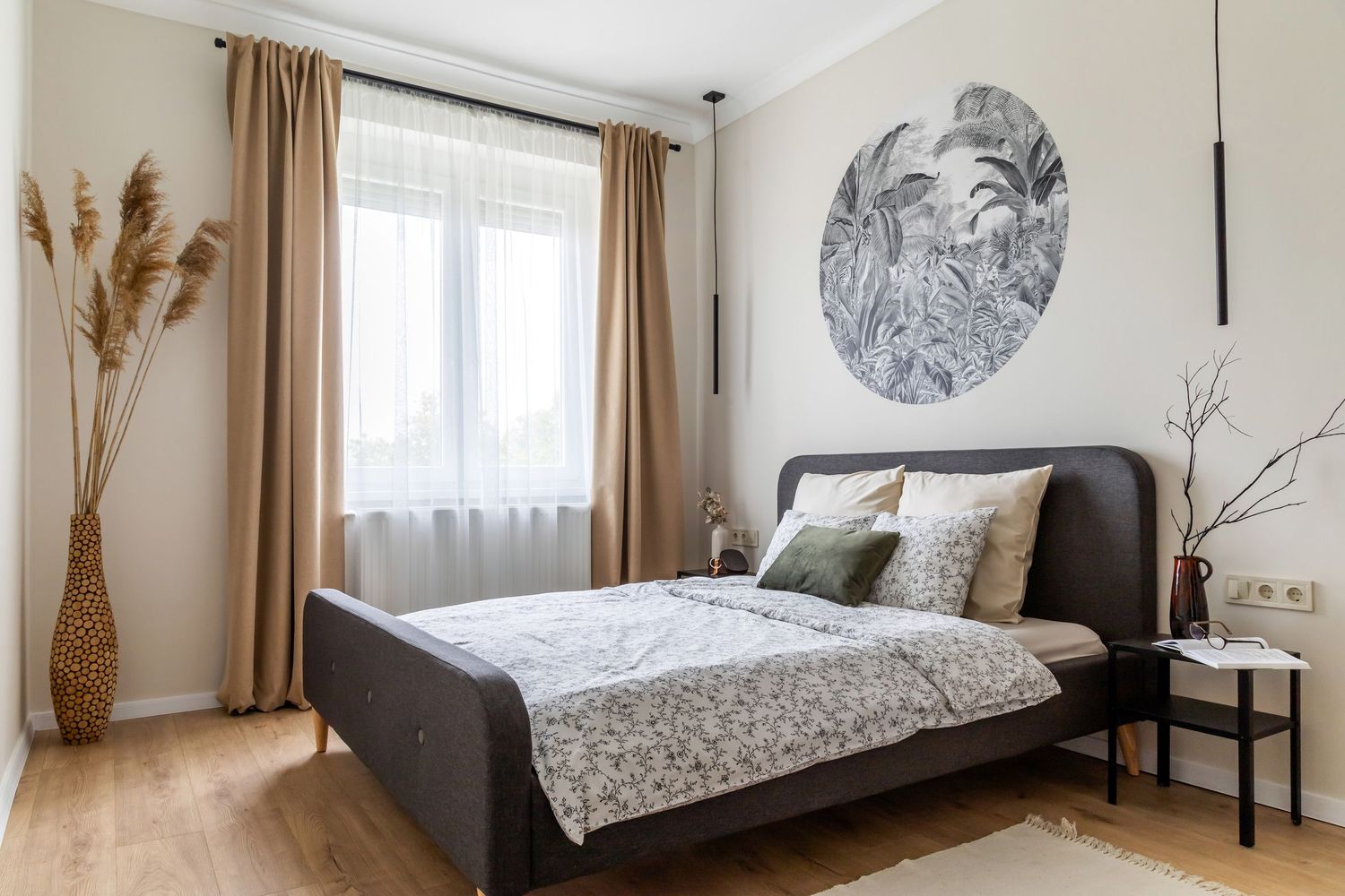 Modern bedroom with dark upholstered bed, botanical wall art, warm beige curtains, and light wood flooring.