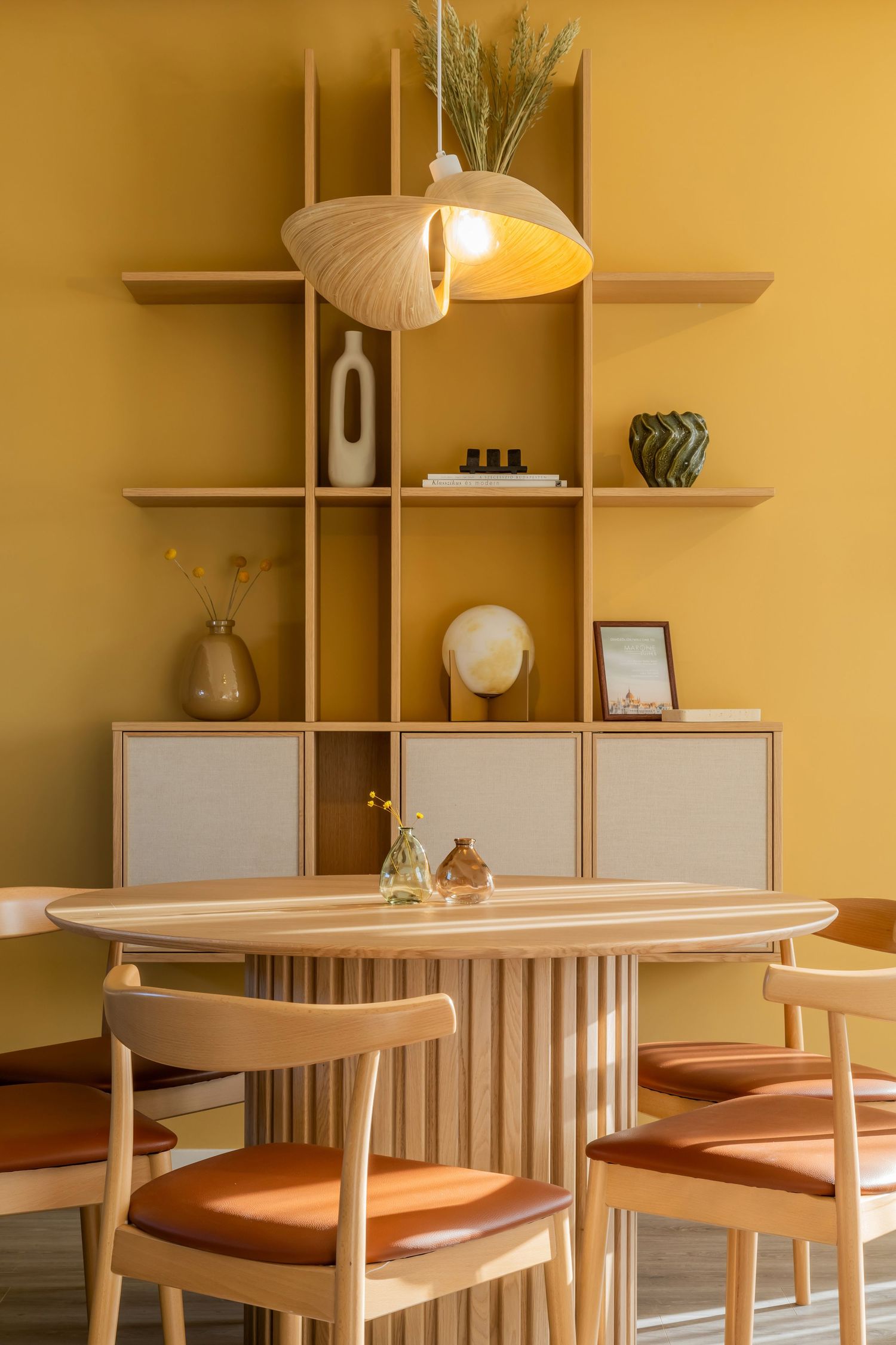 Warm yellow dining room with wooden oval table, leather-seat chairs, pendant lamp, and built-in shelving unit.