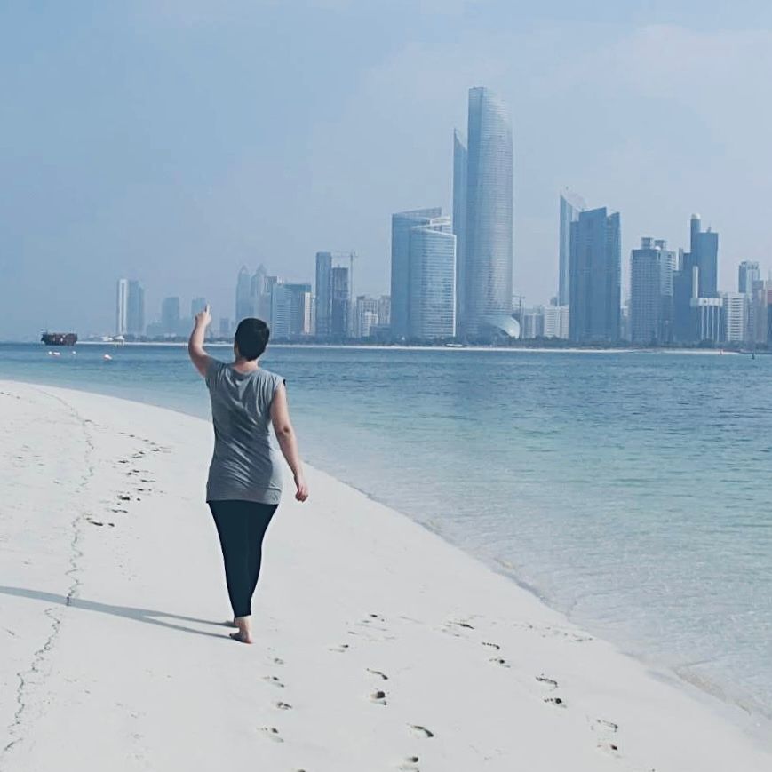 Person walking on white sand beach with city skyline in background, waving hand, clear blue water visible.