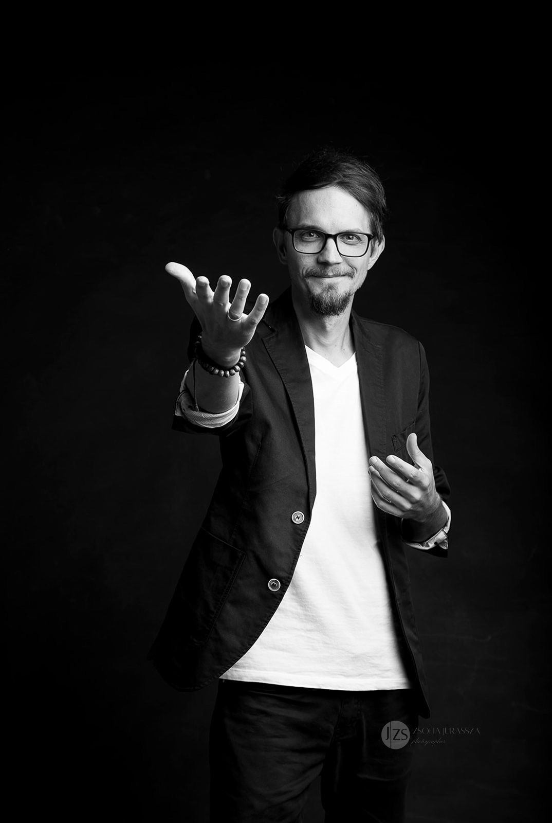 Black and white portrait of a smiling man in blazer reaching toward camera against dark background.