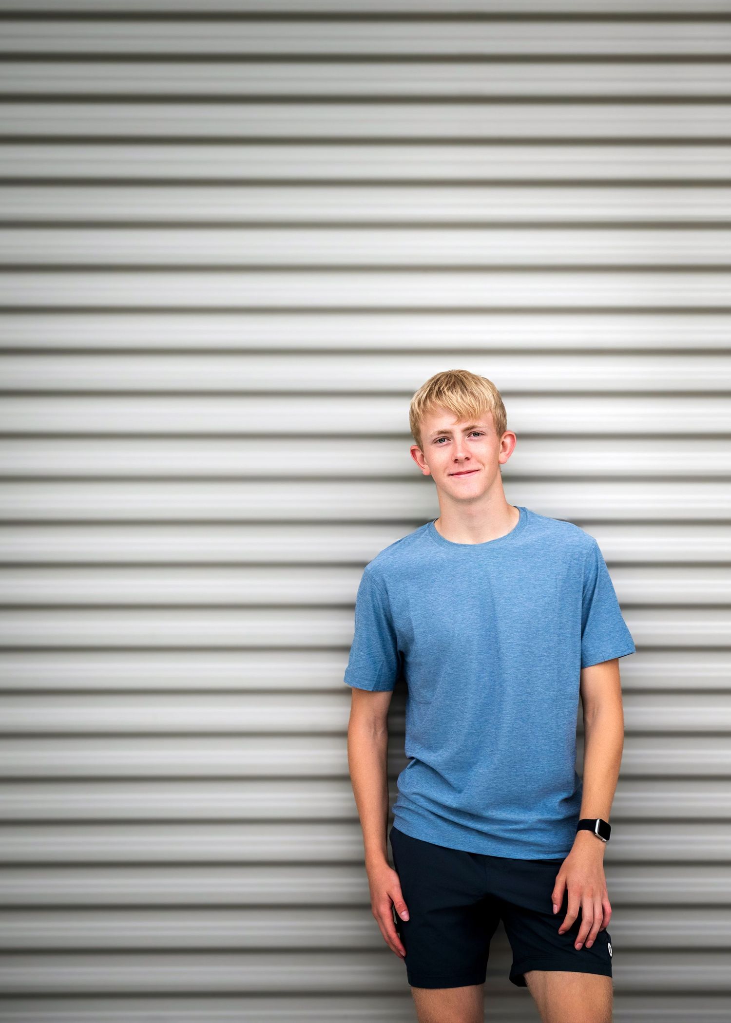 A person wearing a blue t-shirt and dark shorts stands casually in front of a metal roll-up storage door.