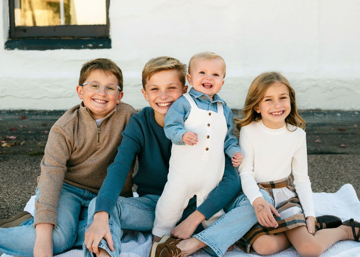 A group of four children sitting together outdoors against a white wall, smiling at the camera.