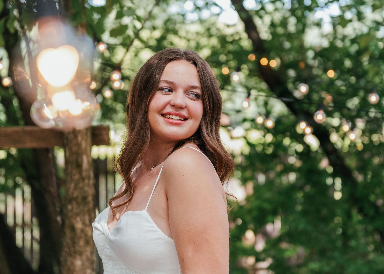 A candid outdoor portrait in a white top with string lights and greenery creating a dreamy summer garden atmosphere.