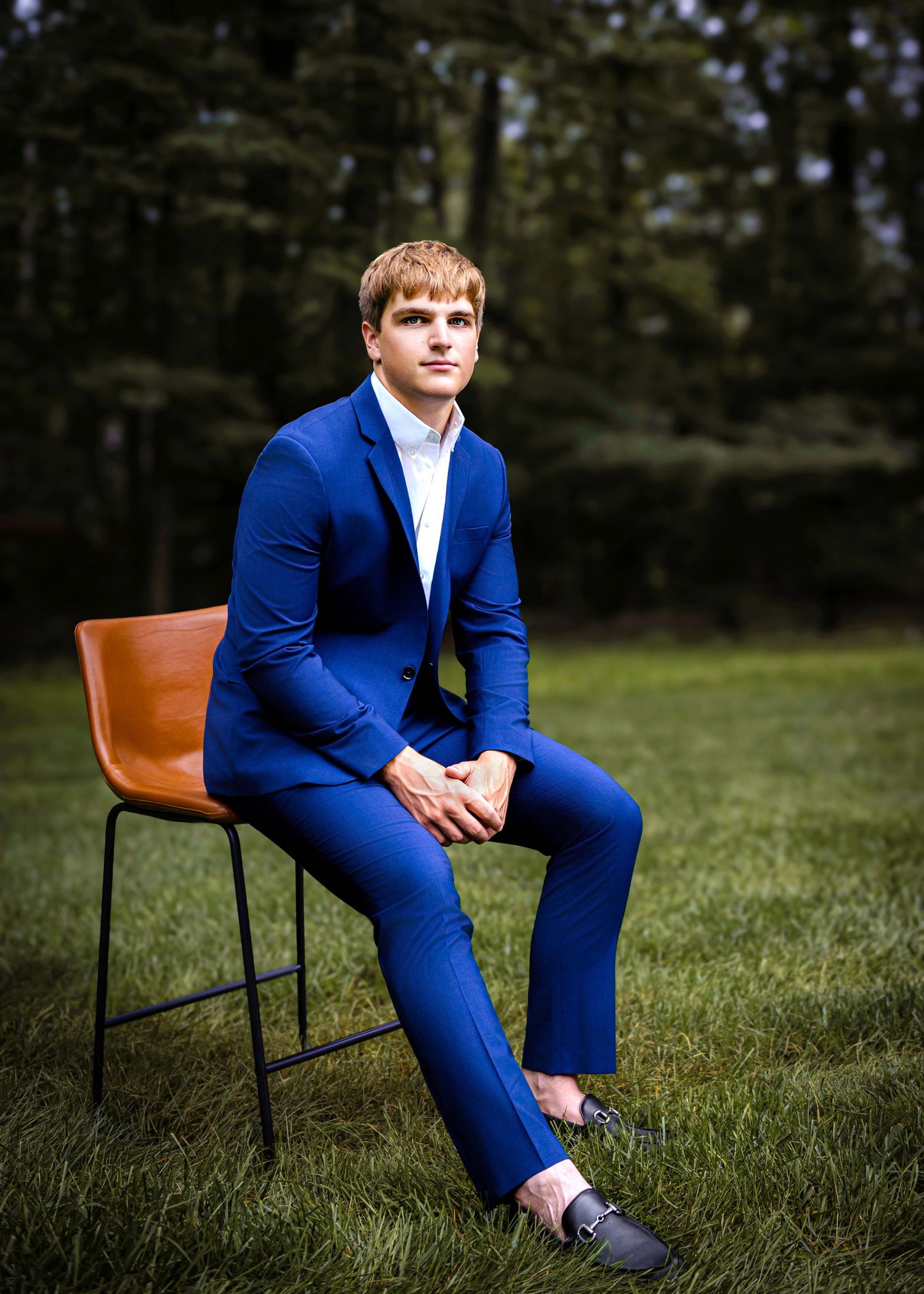 A person in a bright blue suit sits thoughtfully on a wooden chair in a grassy outdoor setting.