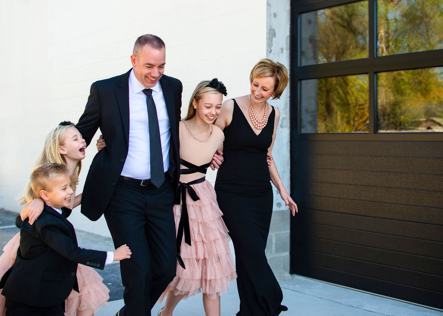 Family group dressed in formal attire laughing together while walking outside a building with glass windows.