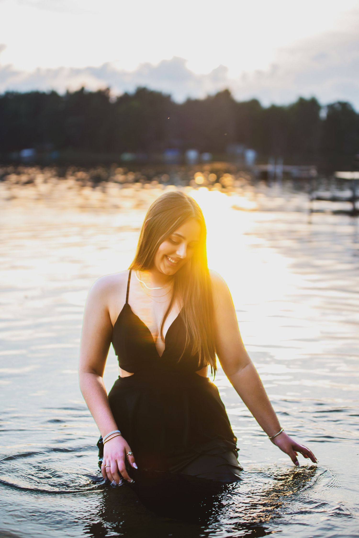 A woman in a black dress sits in shallow water during sunset creating a dreamy, ethereal scene with golden light.