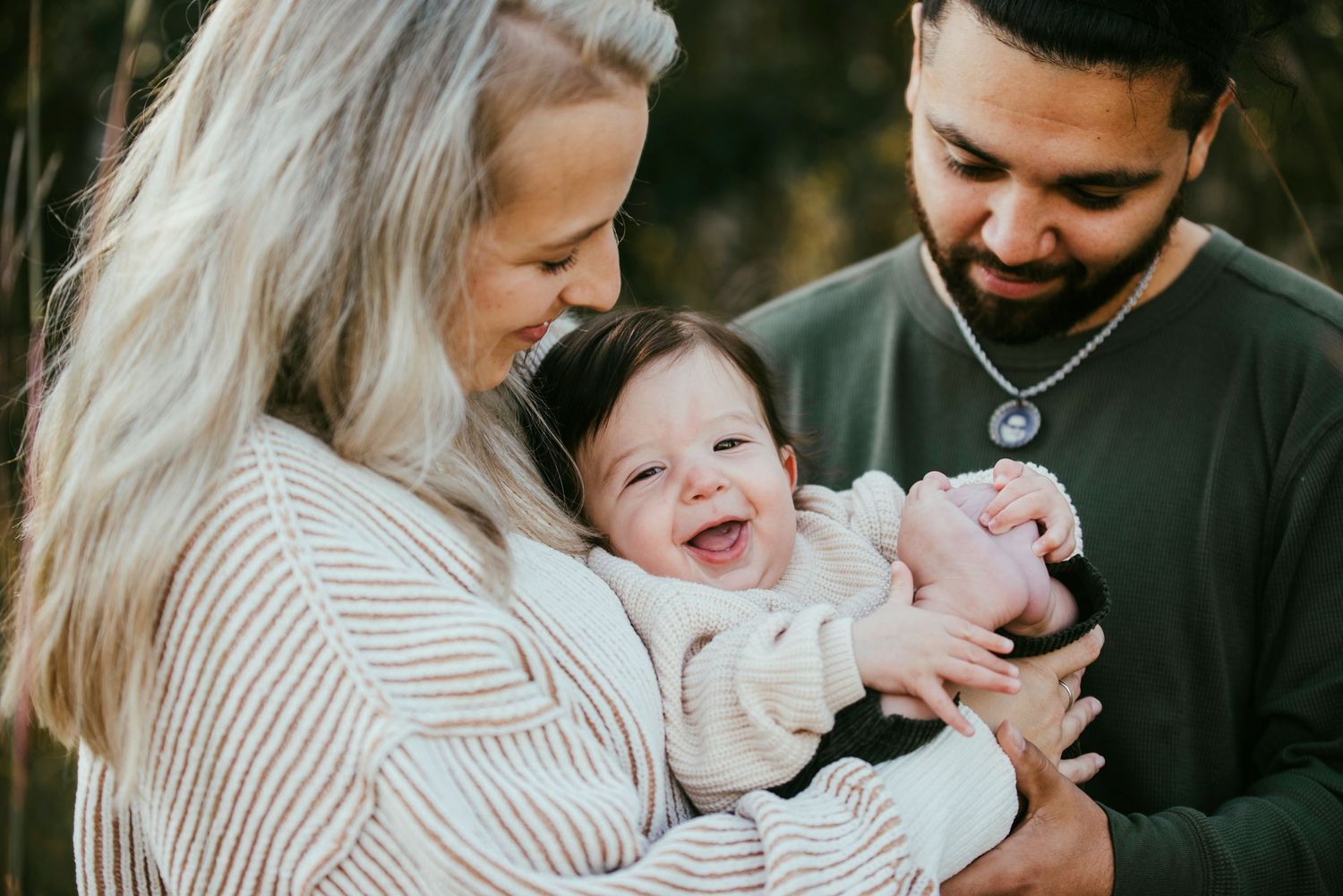 Parents share a joyful moment outdoors with their laughing baby wearing white in natural lighting.