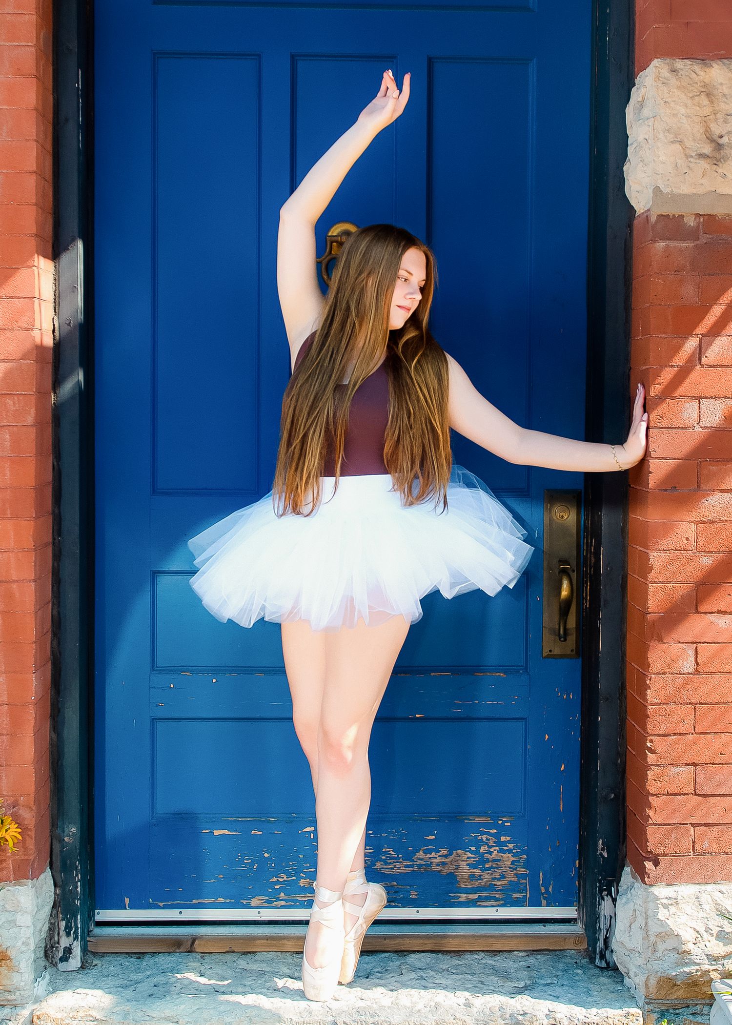 A dancer in a white tutu poses gracefully against a bright blue door and brick wall background.