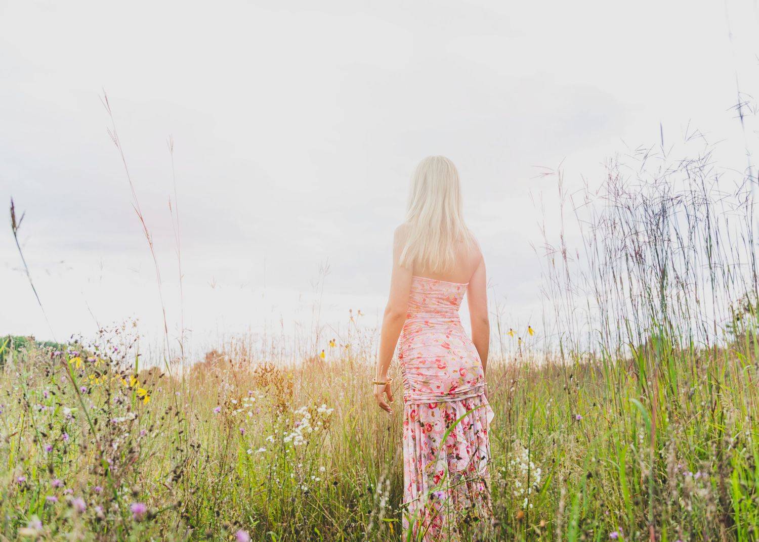 A person in a floral dress stands in a wild summer meadow with tall grass and wildflowers at sunset.