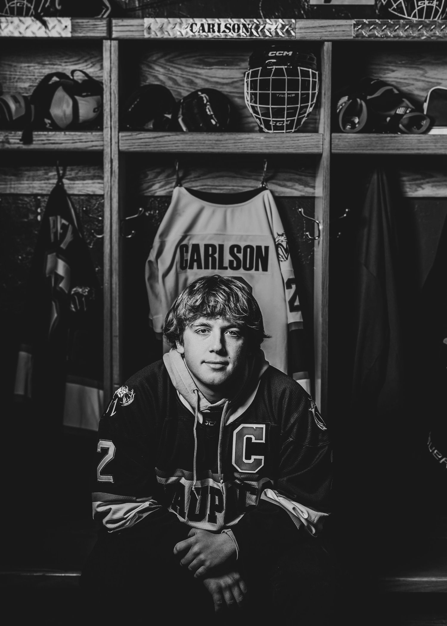 Black and white photo of a hockey player wearing a jersey and  sitting in a locker room stall.