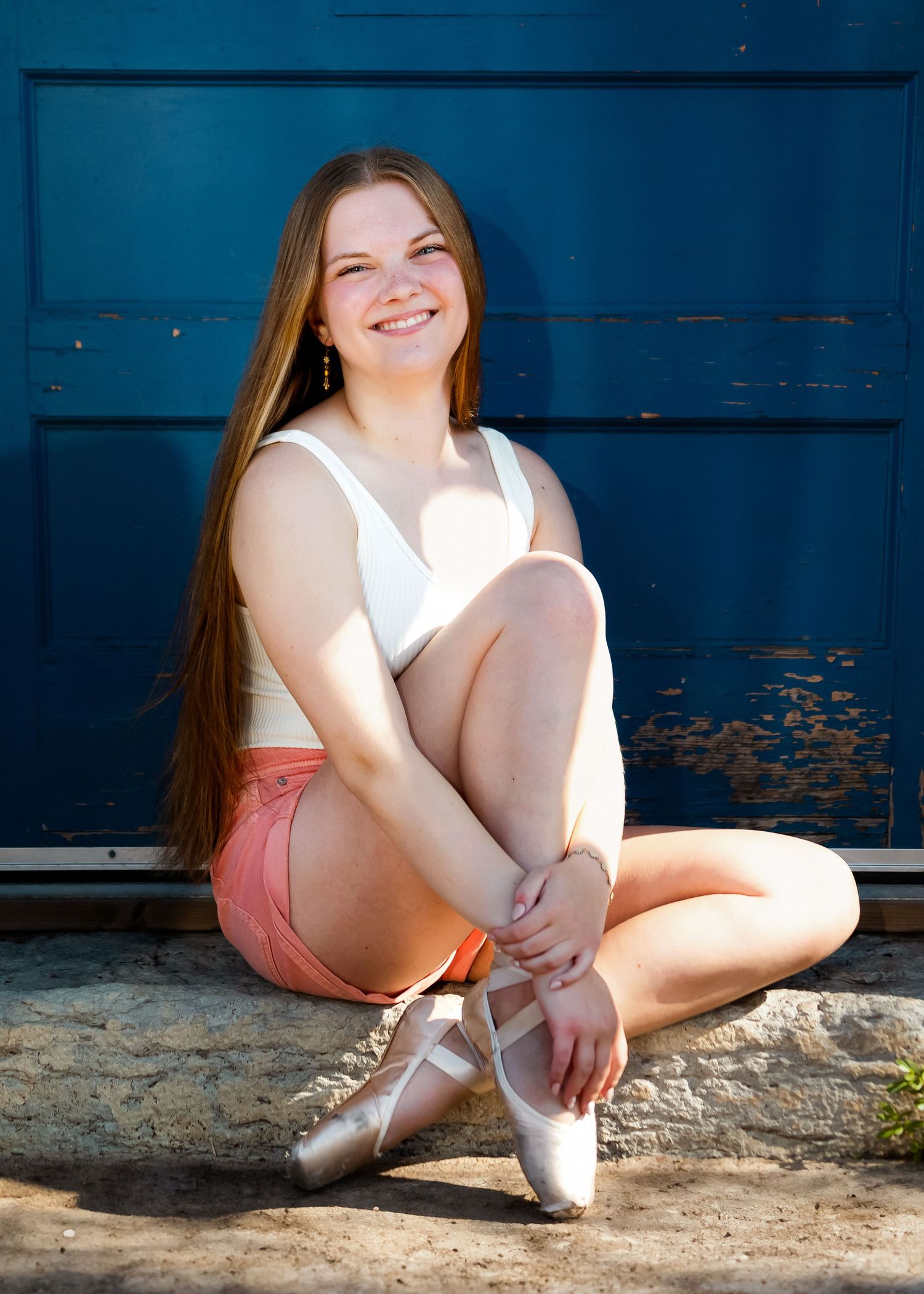 A ballet dancer in pink shorts and a white top sits casually by a blue wall with pointe shoes.