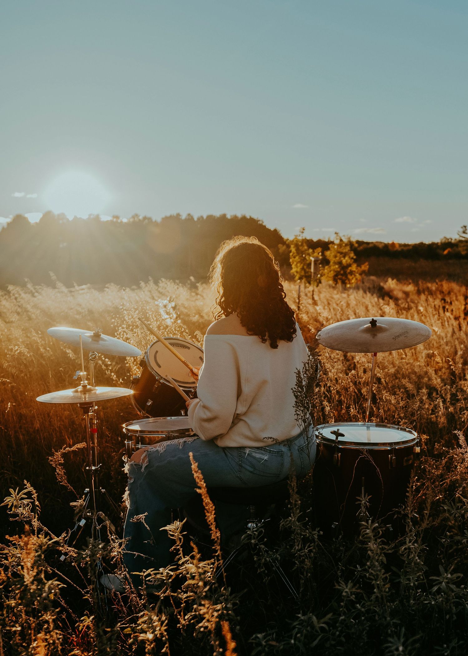 A drummer plays on a drum kit in a golden field during sunset creating a silhouetted artistic scene.