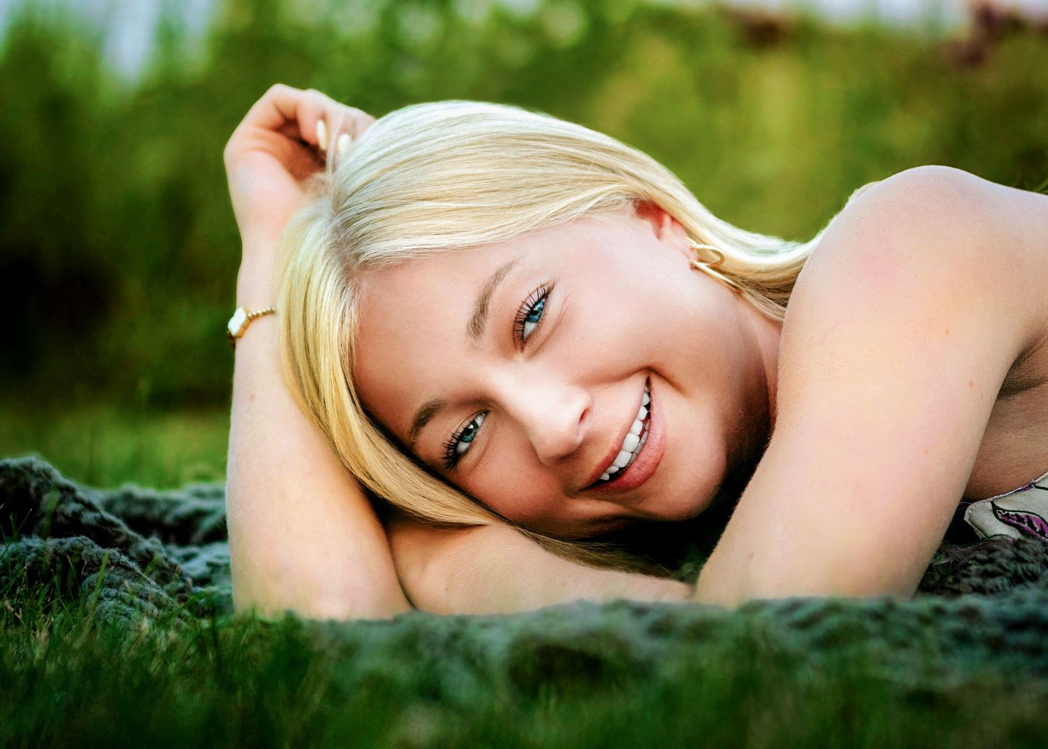 Blonde person lying on grass smiling brightly on a sunny day with green nature in background.
