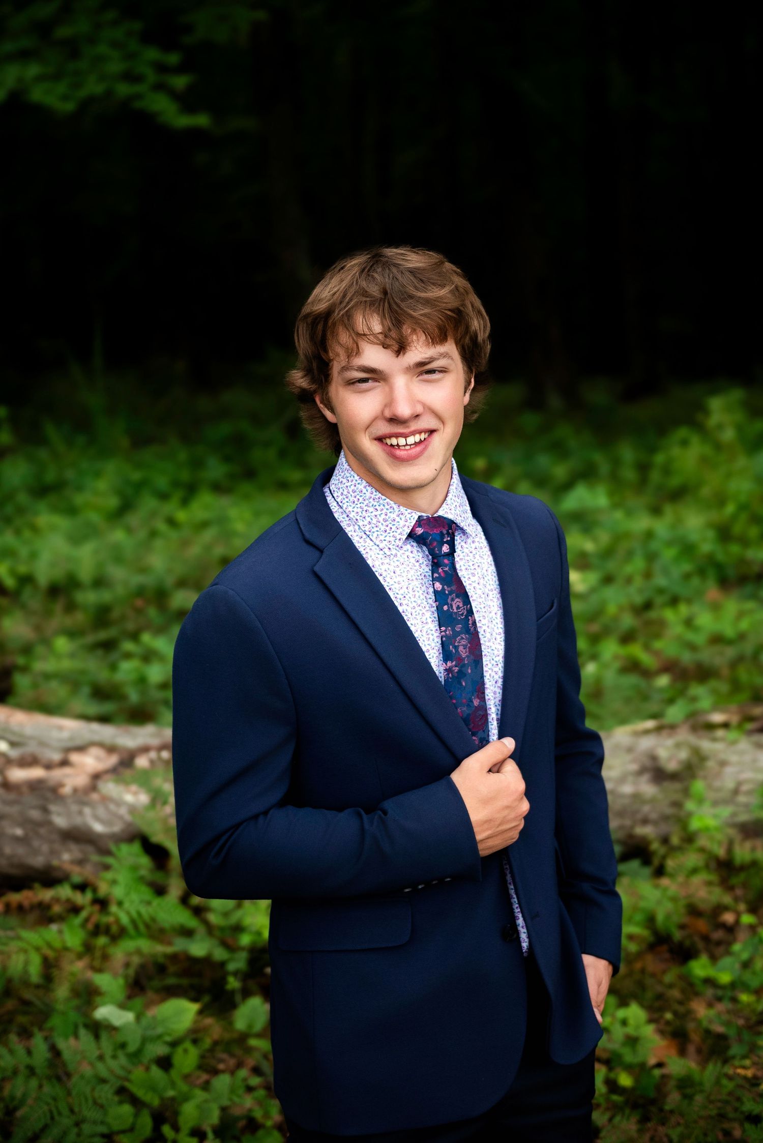 A person wearing a navy suit and polka dot tie stands in front of a natural green outdoor background.