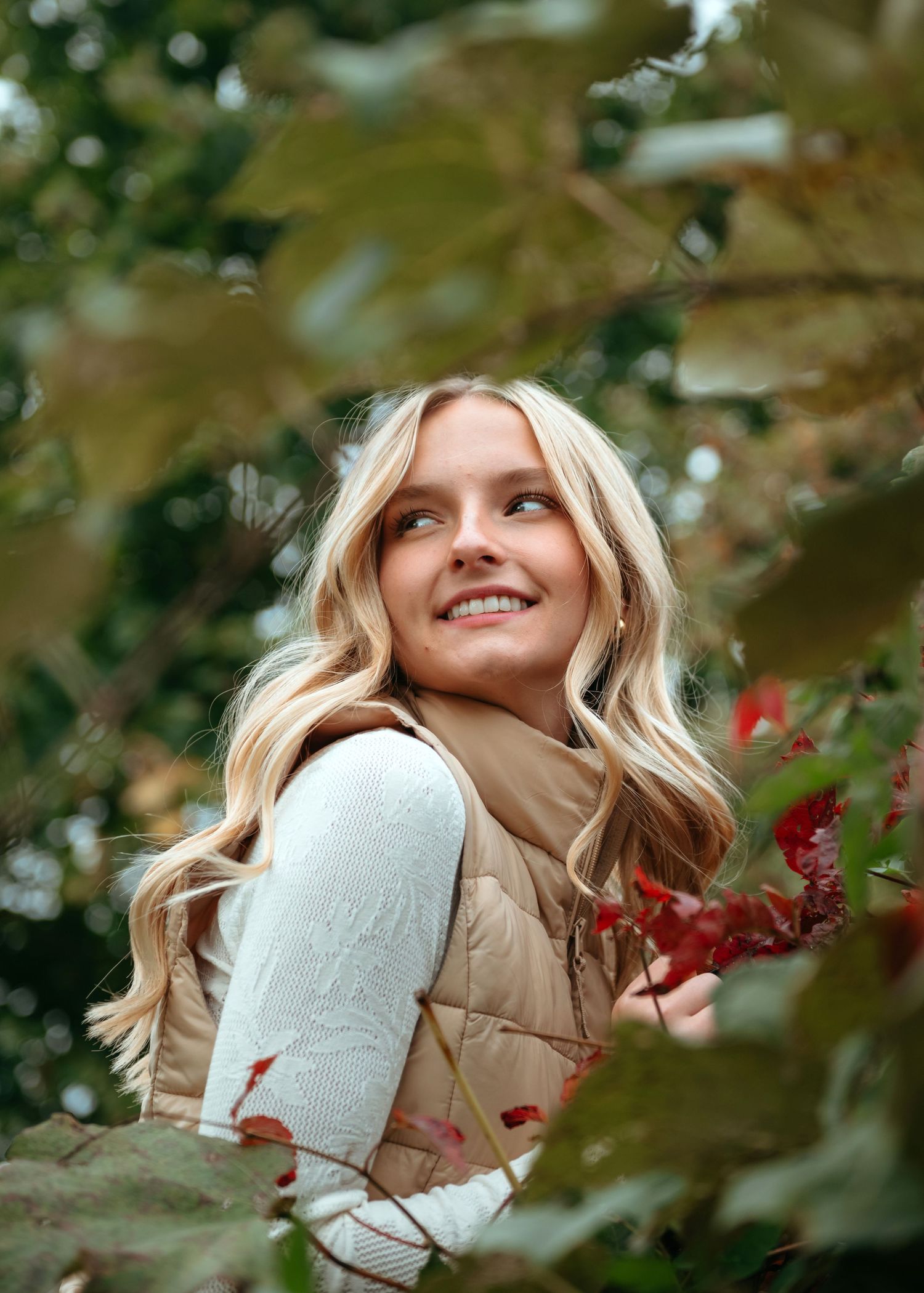A fashion portrait through autumn foliage shows a cozy winter look with a beige coat and mittens among red berries.