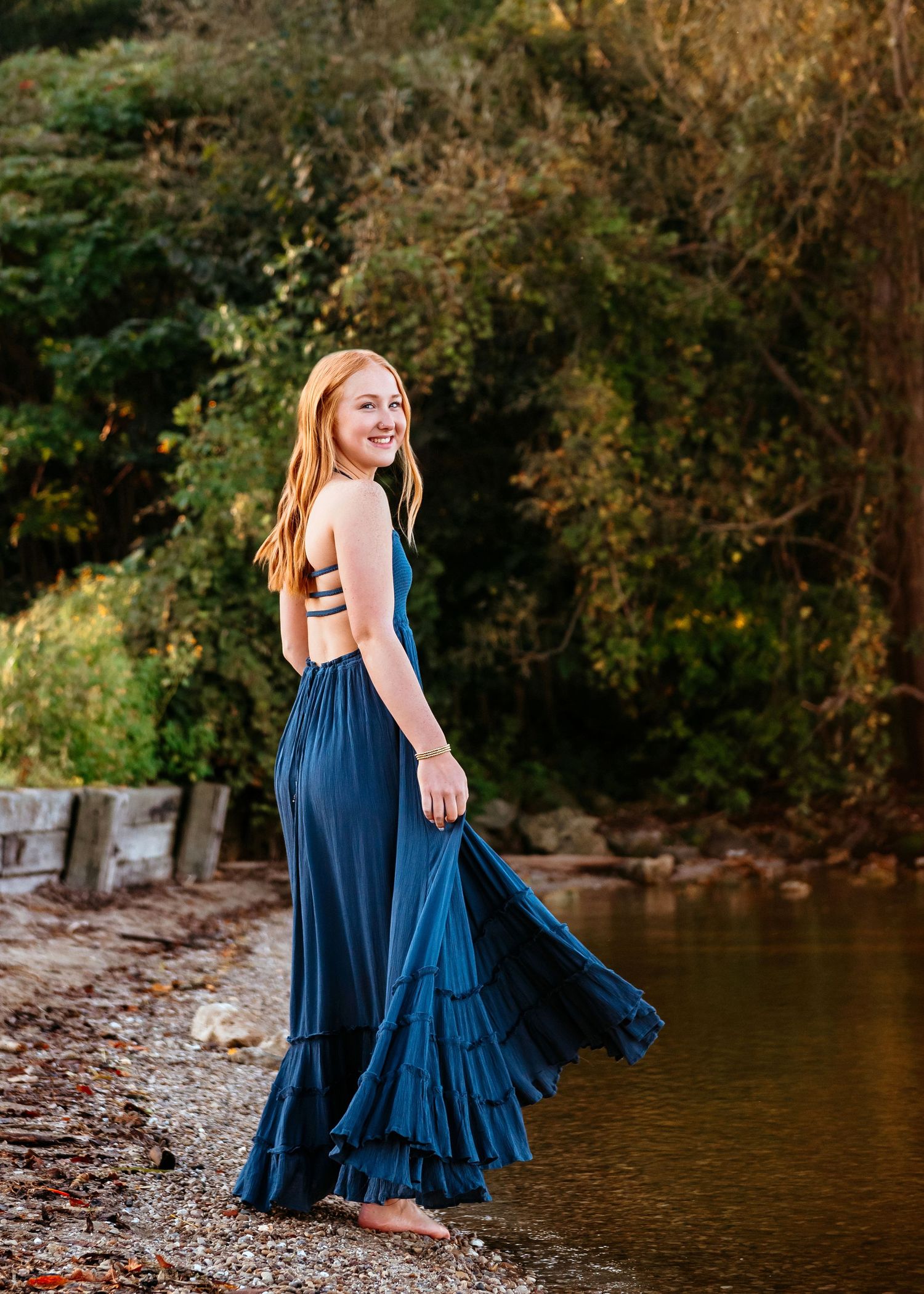 Person in flowing navy blue dress posing by water's edge at sunset.