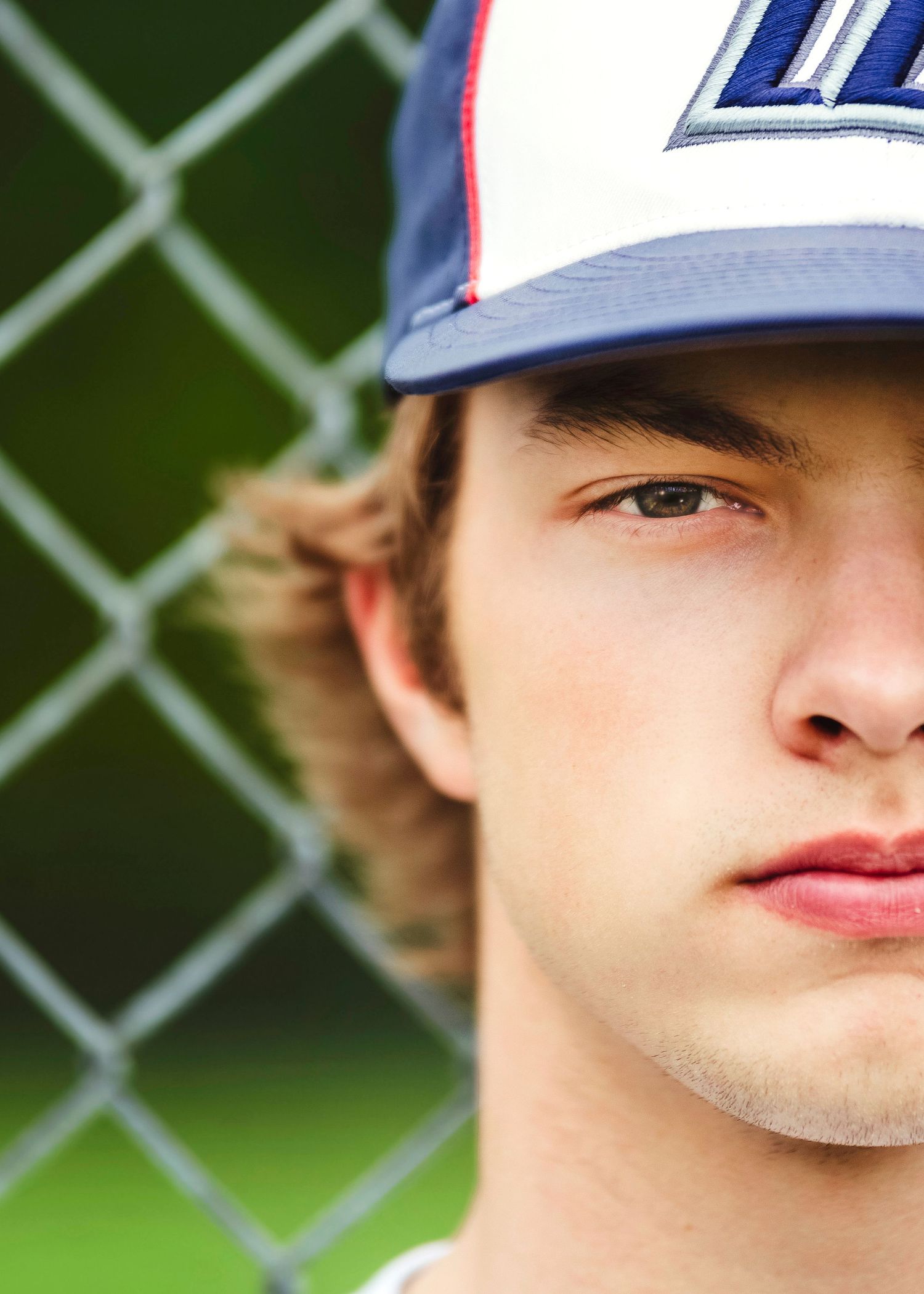 Baseball player wearing a blue and white cap leans against chain link fence on a sunny day.