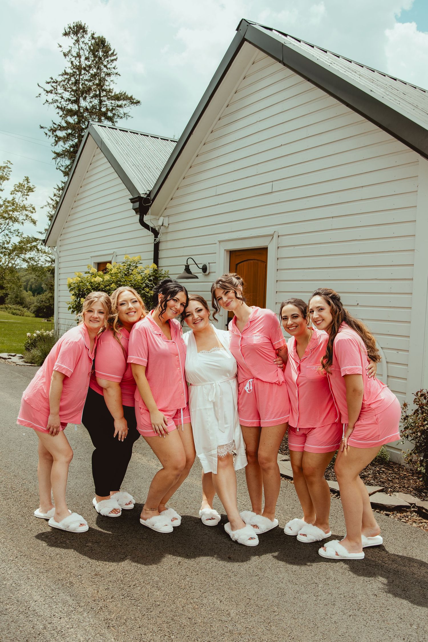 Bride in white robe poses with six bridesmaids wearing matching pink pajama sets and white slippers outside a white barn.