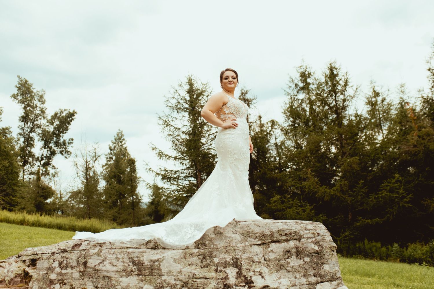 Bride in elegant mermaid wedding gown poses on large rock surrounded by lush green trees.