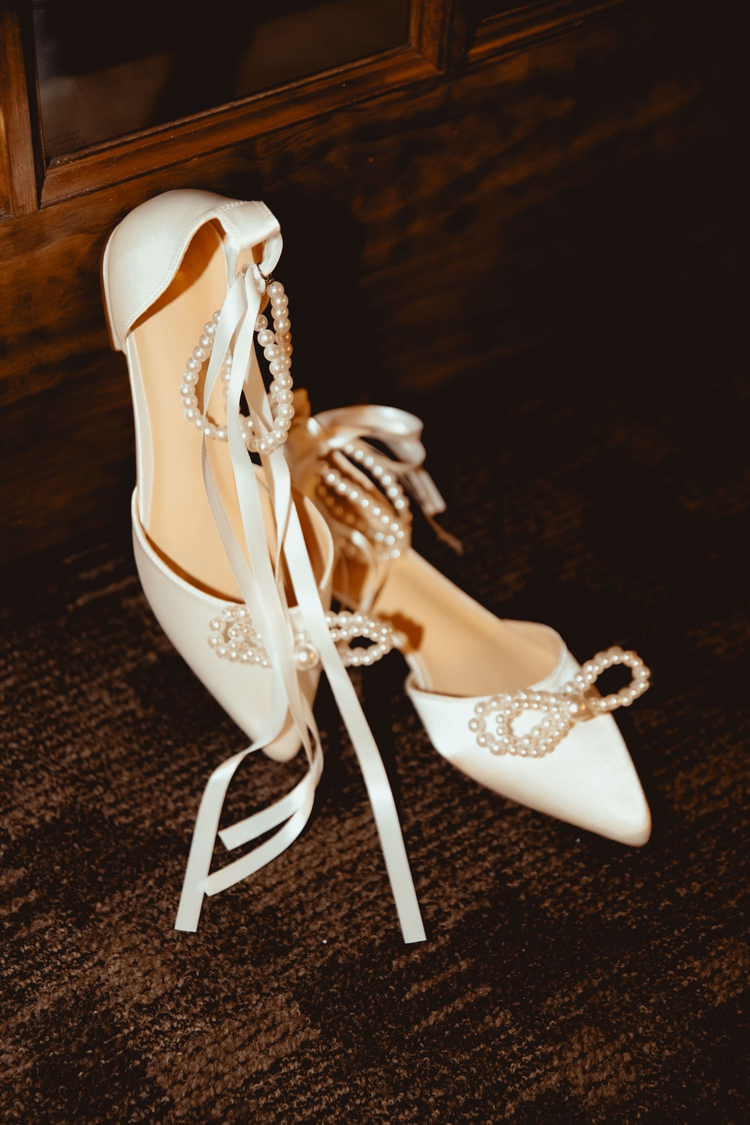 Elegant white satin bridal heels with pearl bow embellishments and ribbon ankle ties on dark carpet.