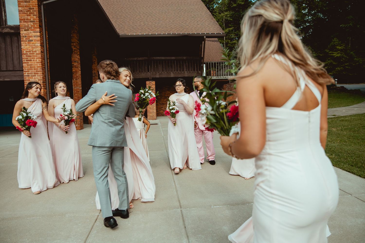 Bride in white gown watches groom embrace bridesmaids in blush pink dresses holding red bouquets outside a chapel.