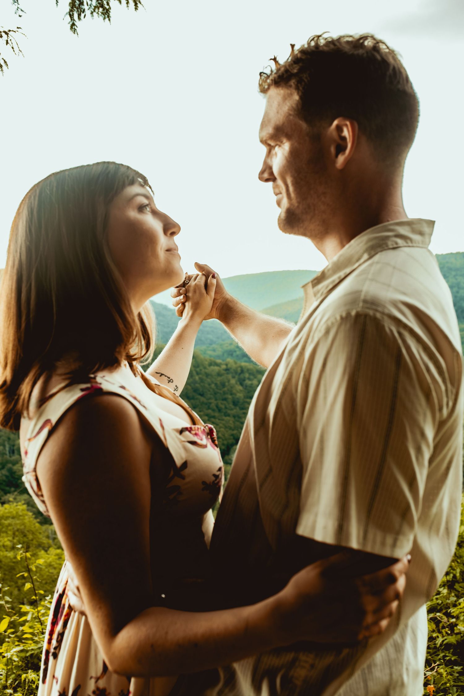 Romantic couple embracing and gazing into each other eyes with sunlit mountain landscape behind them.