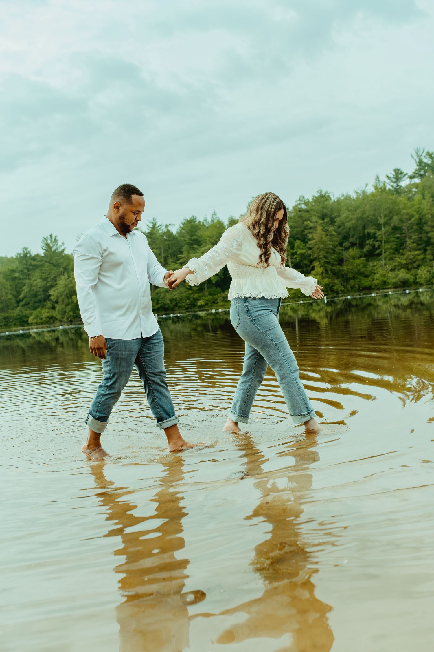 Couple holding hands walking barefoot in shallow lake water with lush green trees in background.