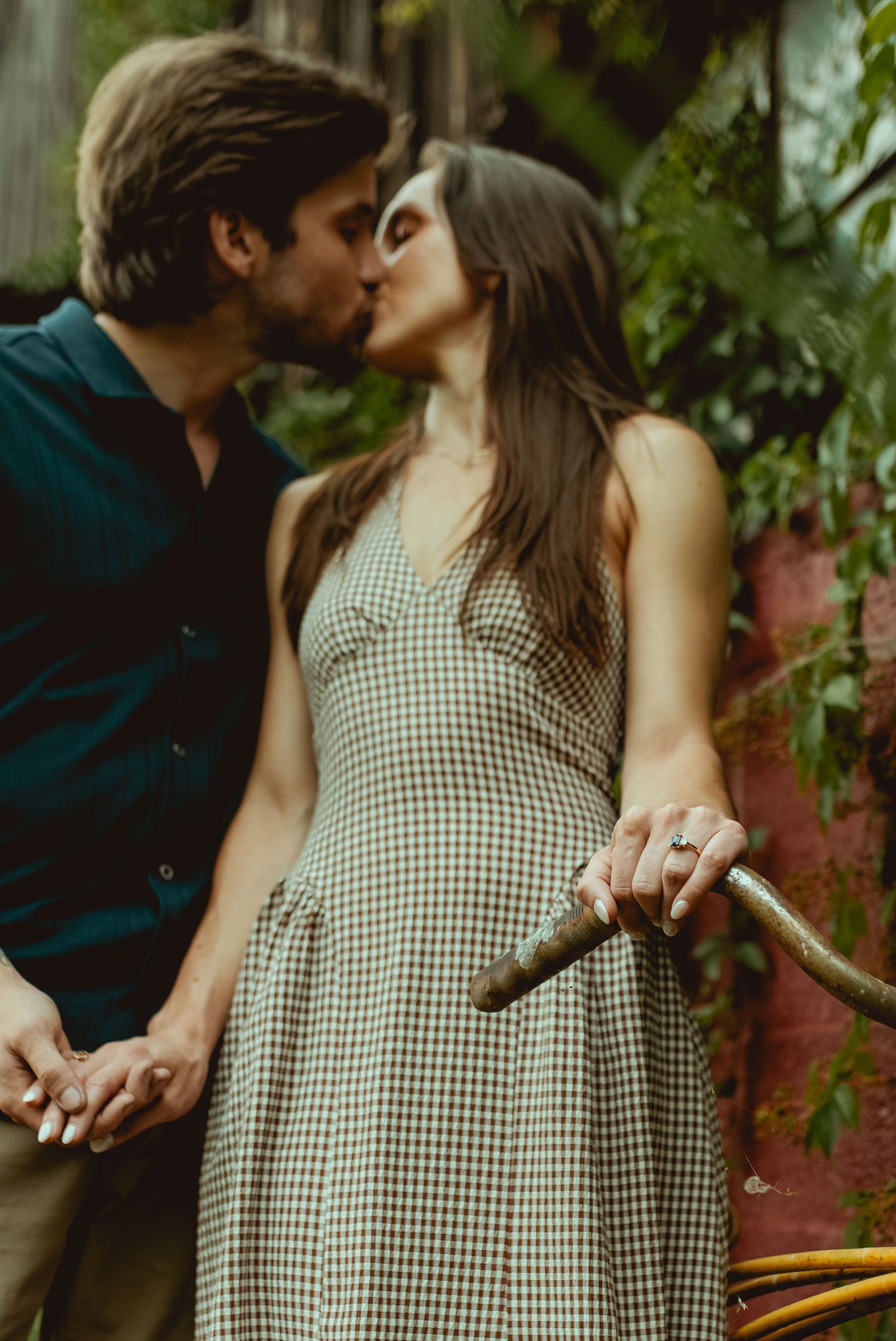 A couple shares a romantic kiss outdoors, woman in checkered dress holding a bicycle, surrounded by lush greenery.