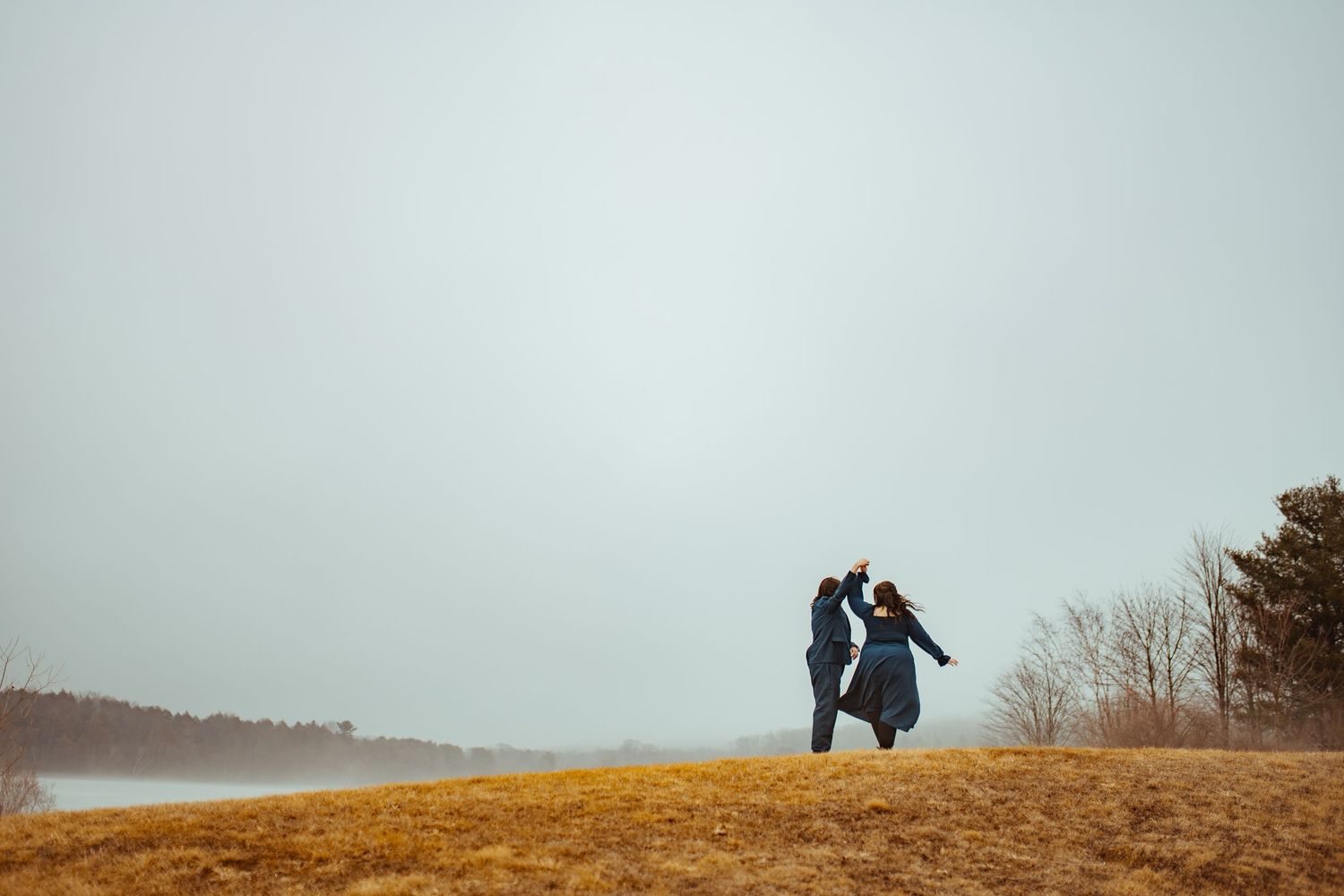 A couple stands silhouetted on a hilltop against an overcast sky during autumn.
