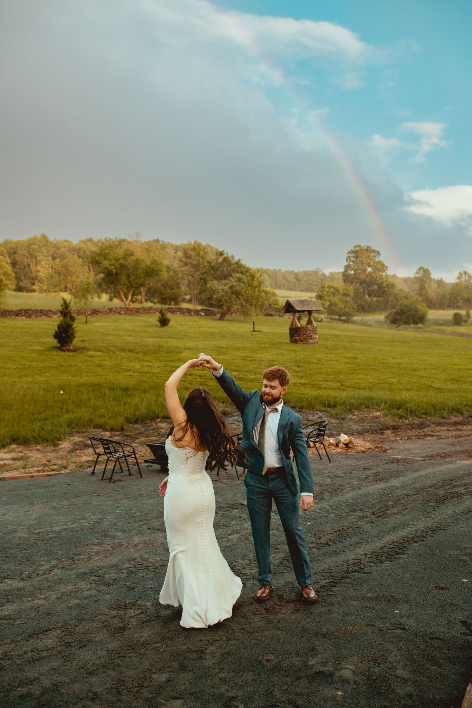 A couple dances together in an open field at sunset with a rainbow visible in the sky.