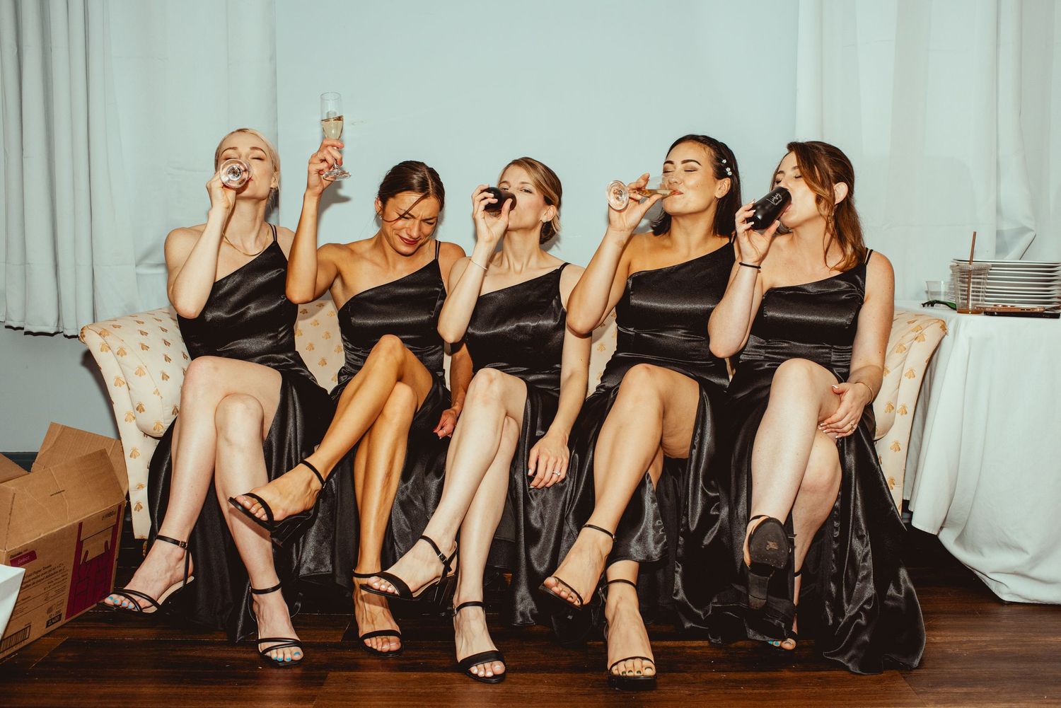 Row of women's feet in formal shoes sitting on a couch during wedding preparations.