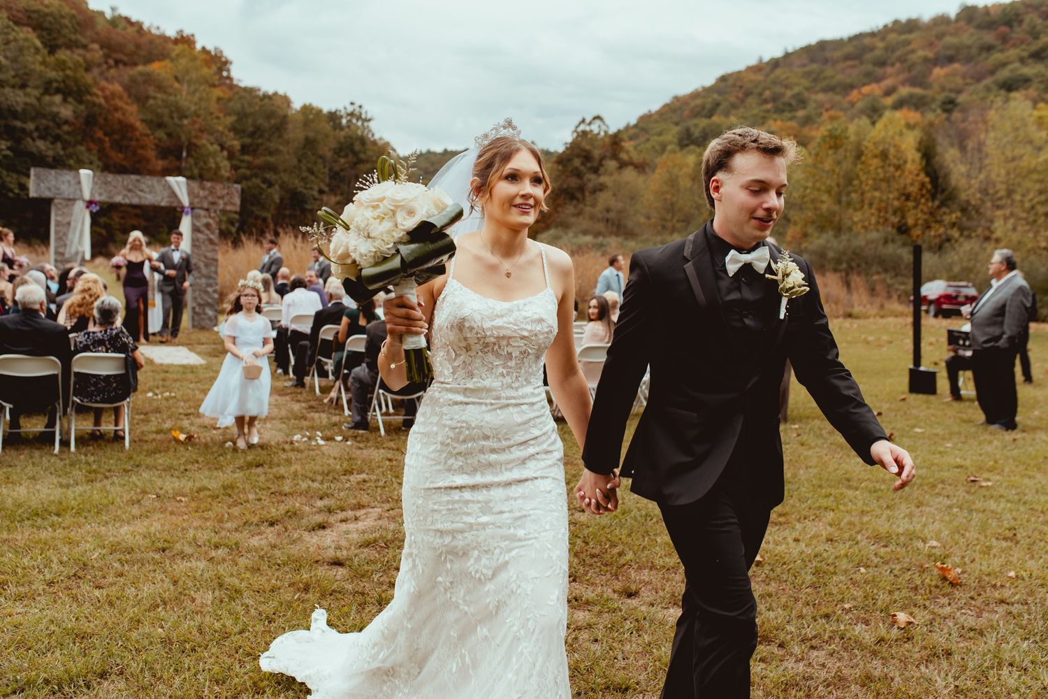 A newlywed couple walks hand in hand across a grassy field with autumn hills in the background.