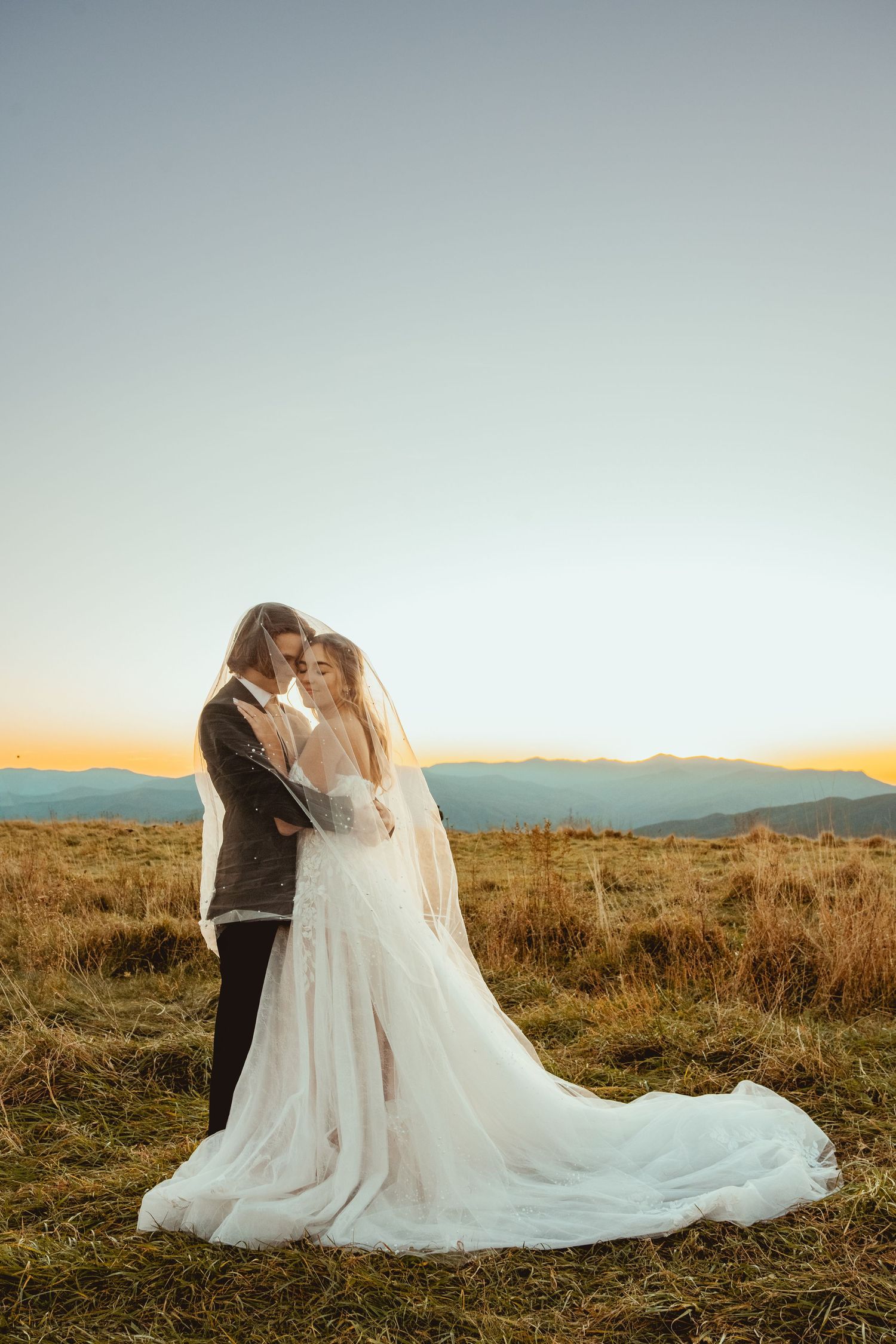 A wedding couple embraces on a hilltop with mountains in the distance at sunset.