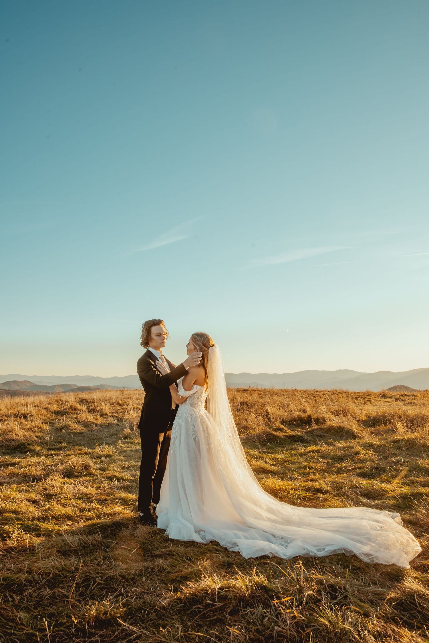 Newlywed couple embracing in golden sunlit field with mountains in background.