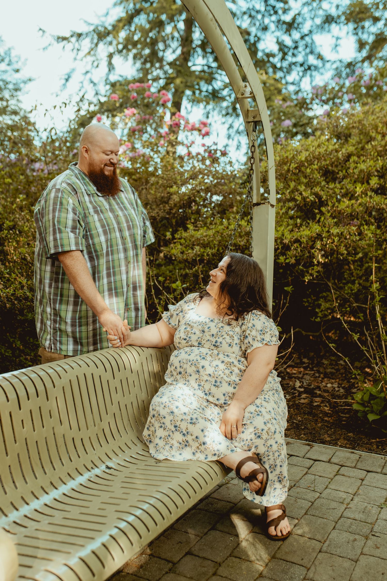 A couple holds hands on a garden bench, the woman in a floral dress smiling up at her bearded partner outdoors.